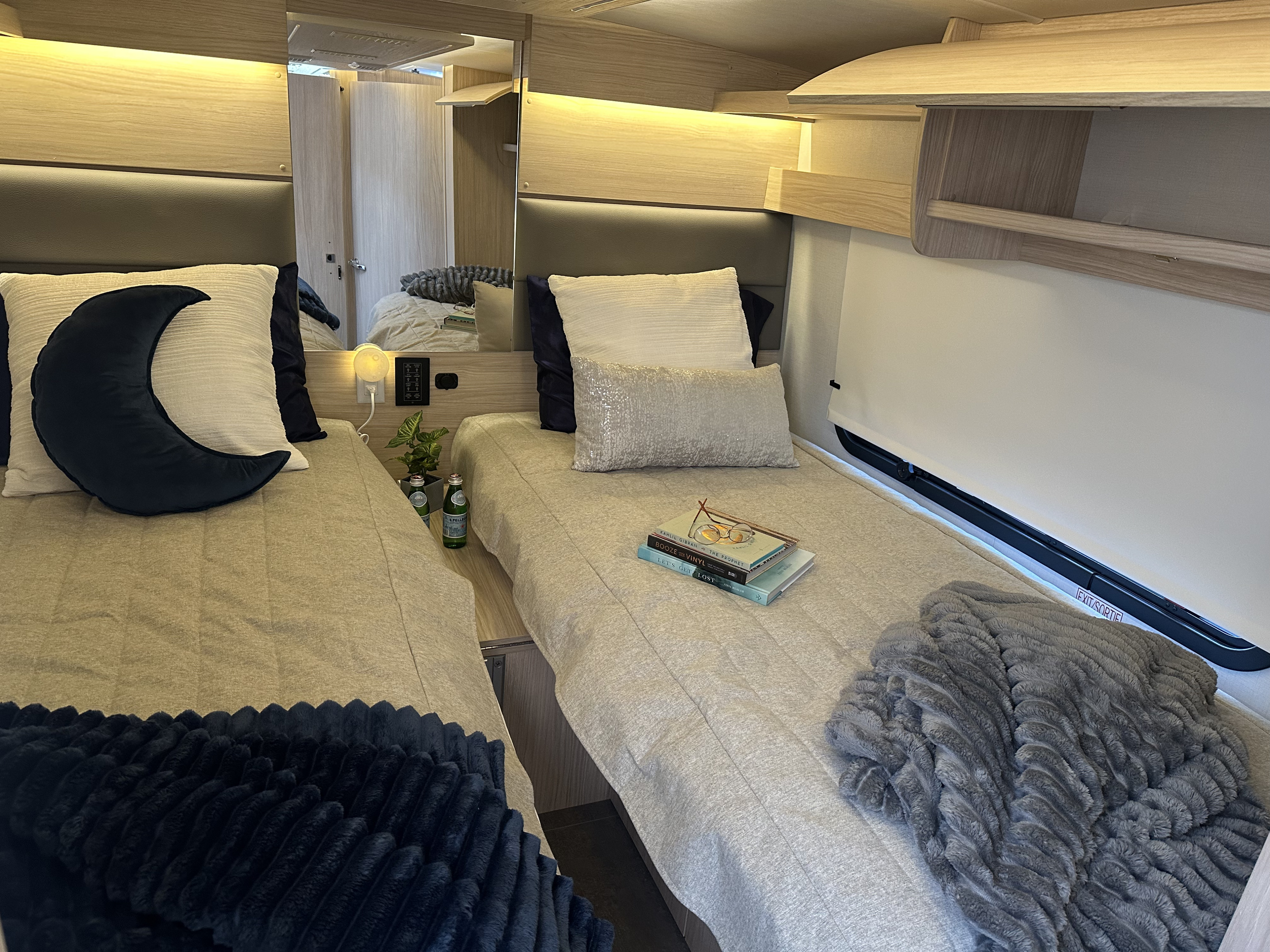 Comfortable RV rental bedroom featuring twin beds with moon pillow, books, and cozy blankets, framed by wooden cabinetry.. Leisure Travel Van Wonder 24RTB 2025