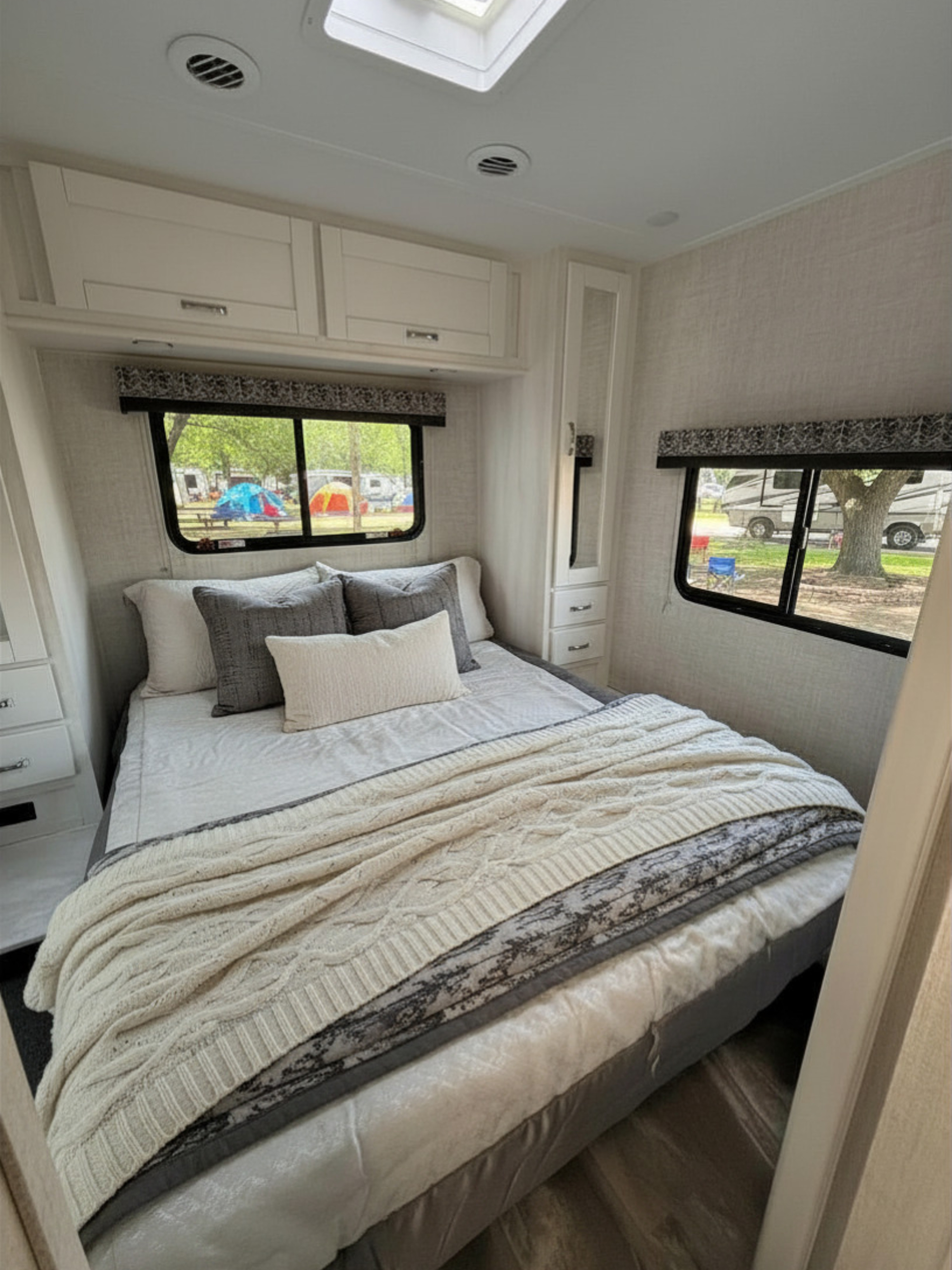 Cozy RV rental bedroom features a comfy bed with a cable knit throw, overhead cabinets, and a bright campsite view.. Gulf Stream Conquest 2022