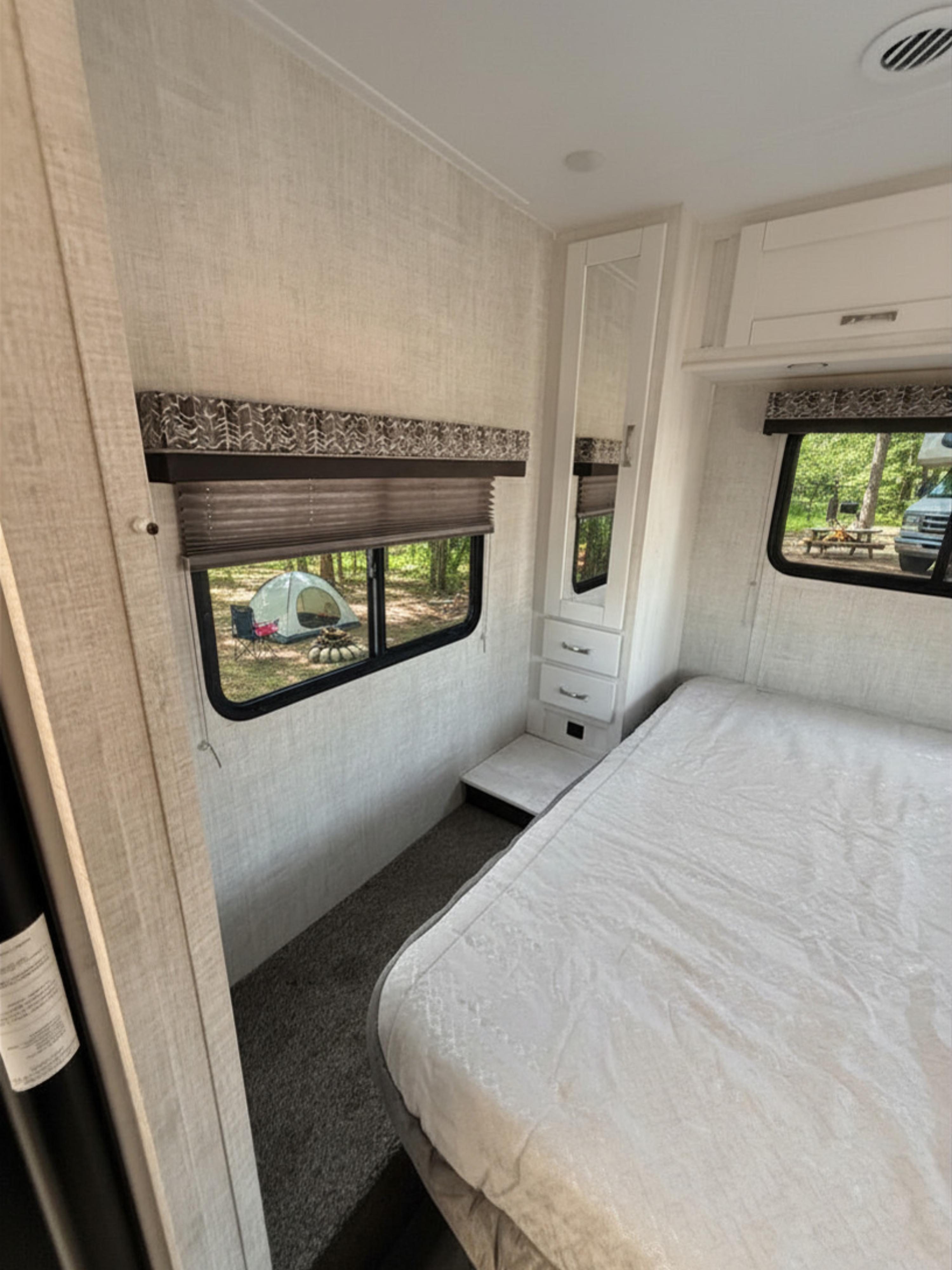 An RV rental bedroom with white bedding & cabinets. Windows show a campsite with a tent, fire pit, and trees outside.. Gulf Stream Conquest 2022