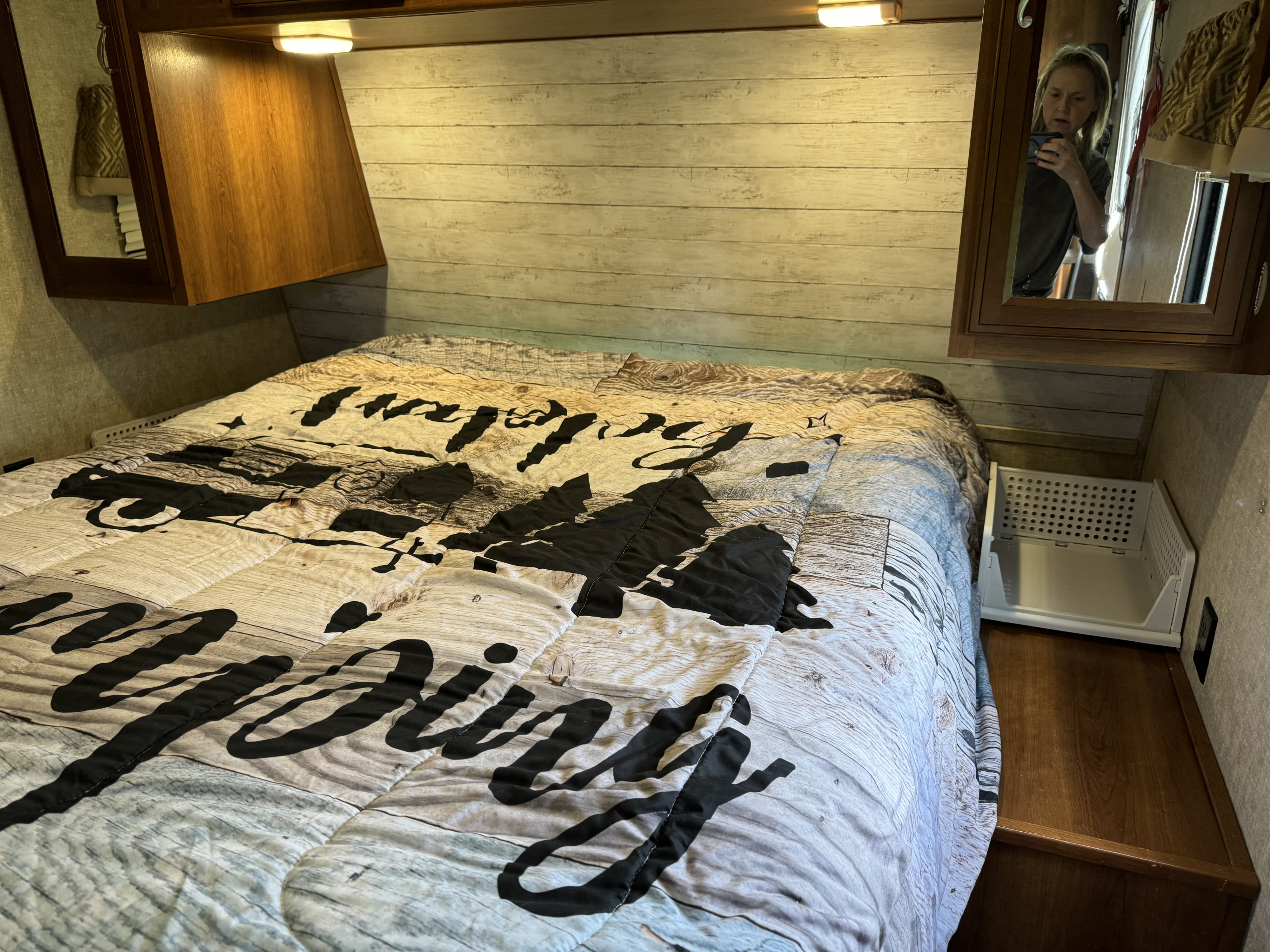 Cozy RV rental bedroom with a rustic wood-print comforter and overhead cabinets. Mirror reflects person taking photo.. Avenger ATI 2017