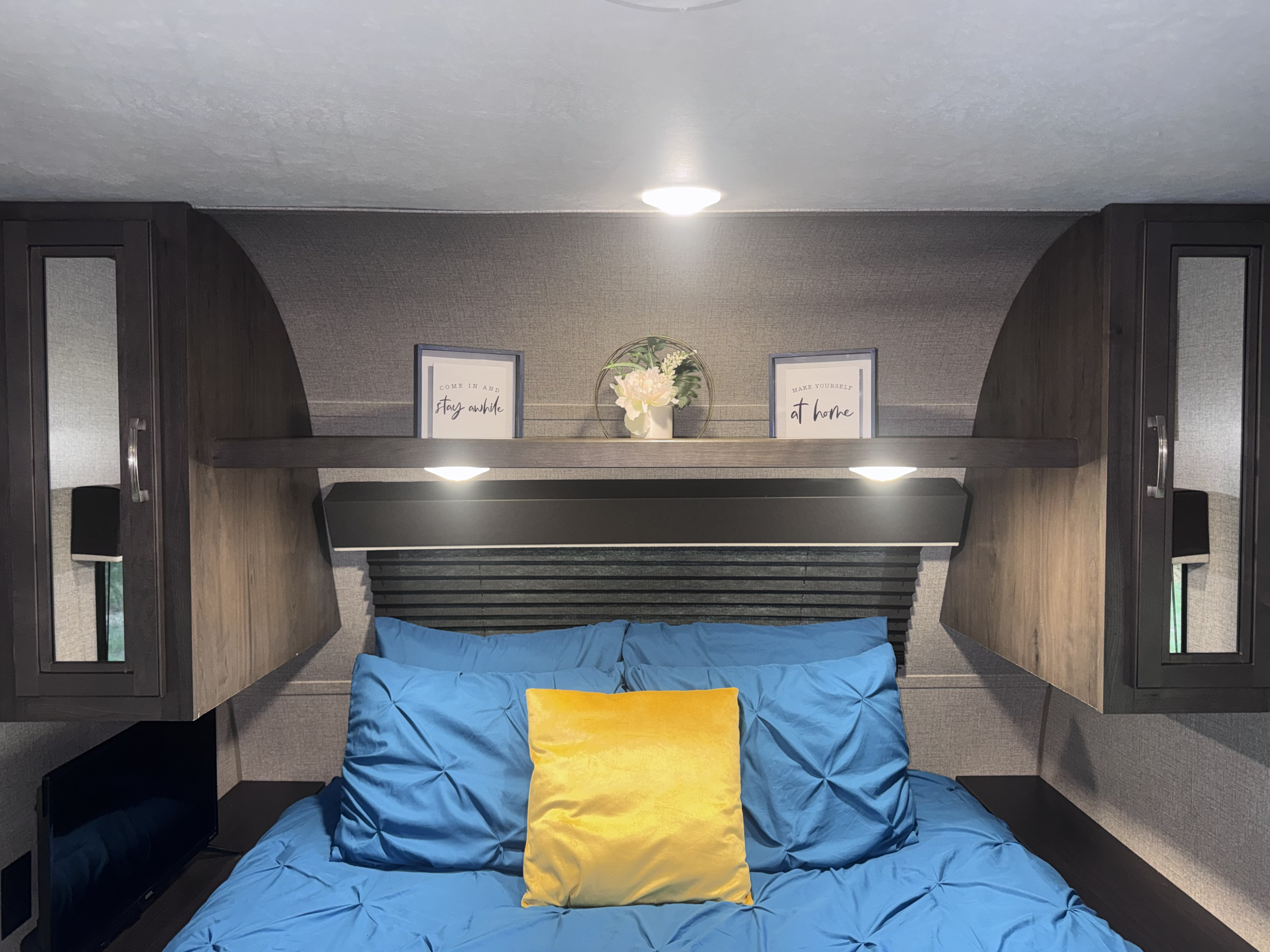 Inside an RV rental: a cozy bedroom featuring a blue bed, yellow pillow, overhead cabinets, and decorative shelf items.. Jayco Jay Flight SLX 2020