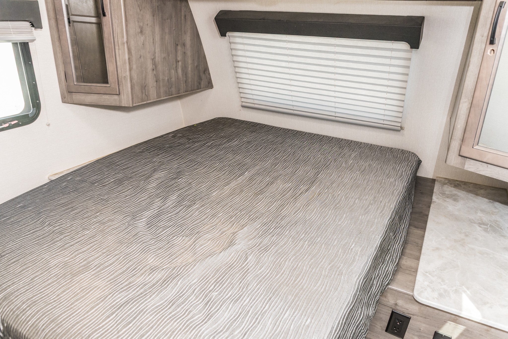 Modern RV rental bedroom: gray textured bed, wood cabinet, window blinds, and counter space.. Coachmen Apex 2021