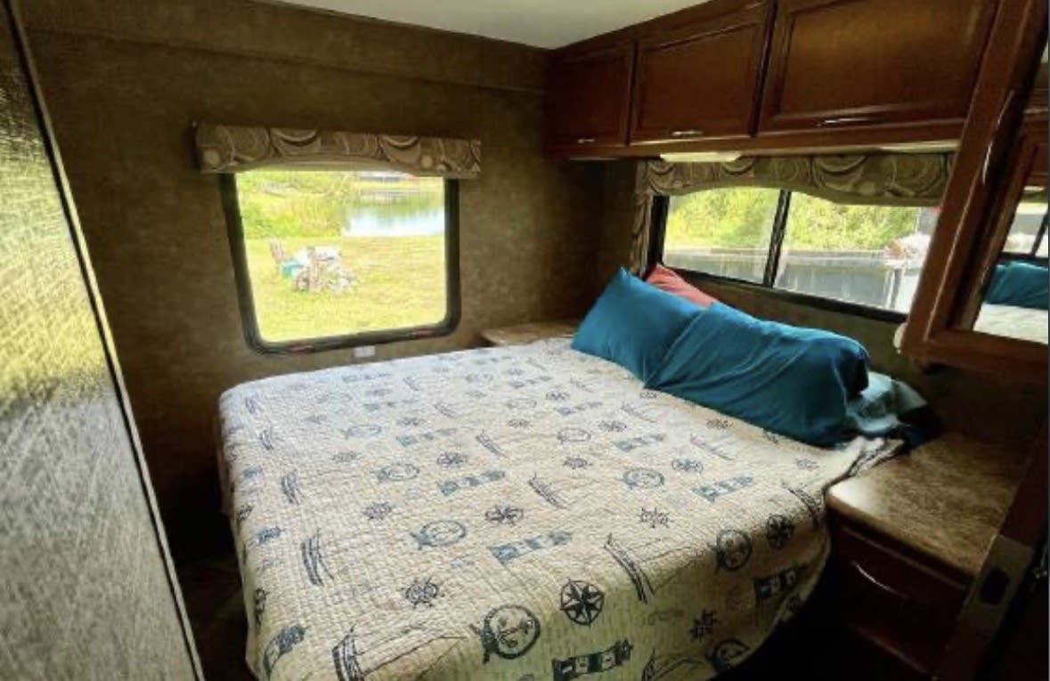 RV rental bedroom: cozy bed with nautical quilt, blue pillows, and a window view of a grassy area and lake from inside the camper.. Thor Four Winds 2014