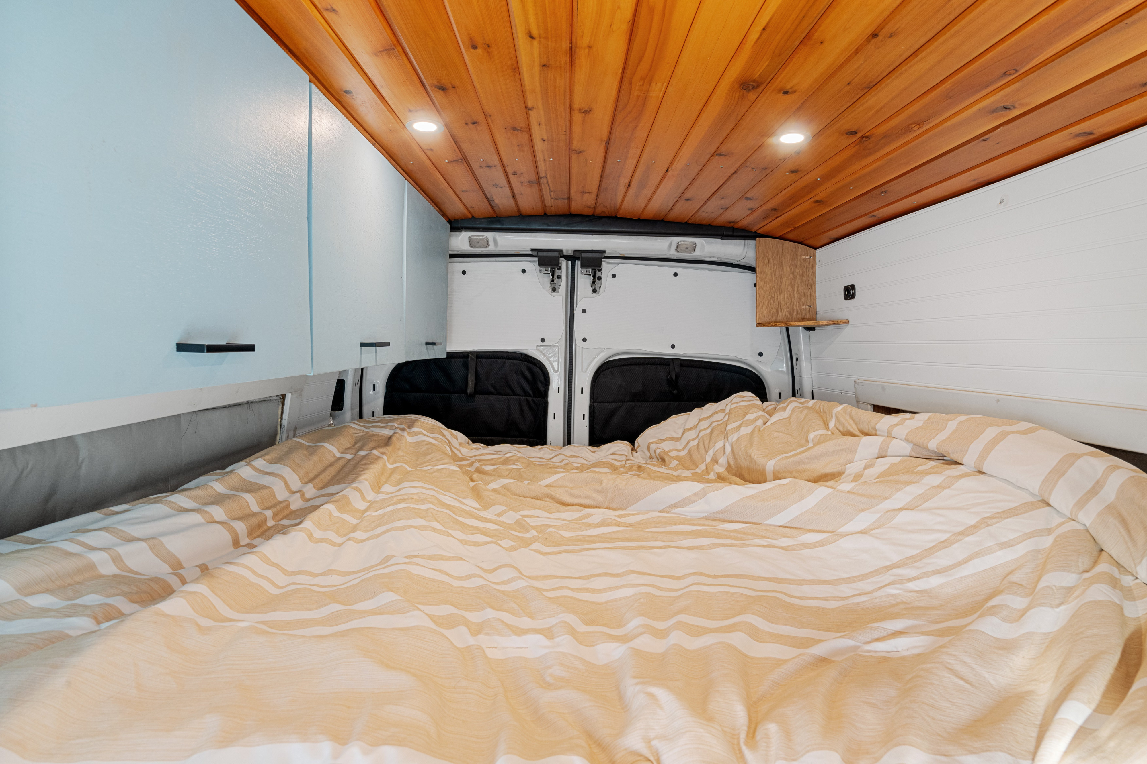 Relaxing rv rental bedroom featuring striped bedding, wooden ceiling with lights, and light blue cabinetry.. Ford Van 2015