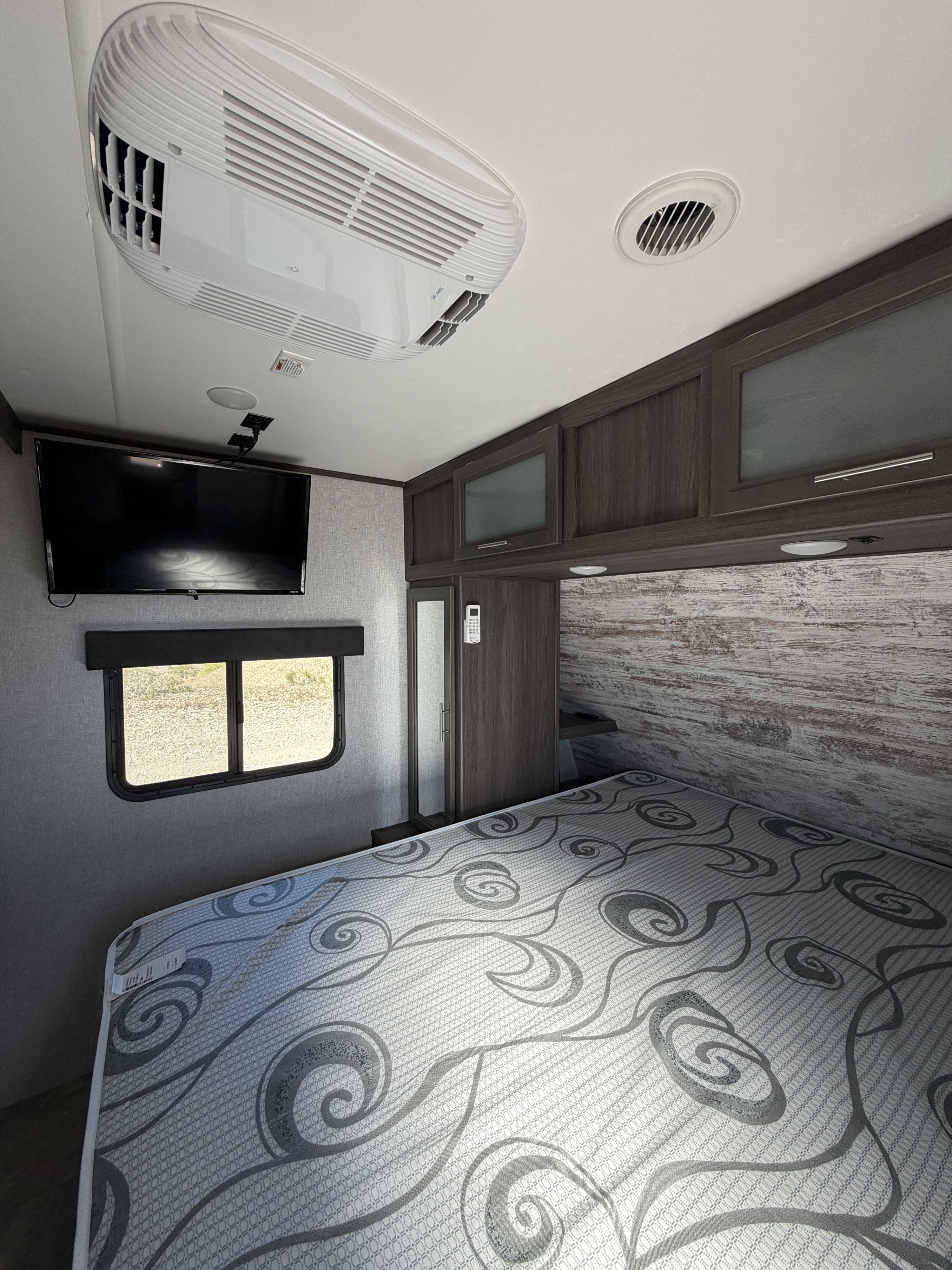 RV rental bedroom with patterned mattress, RV AC unit, TV, and wood-look cabinets. Cozy, modern travel trailer interior.. Dutchmen Kodiak Ultra Lite 2021