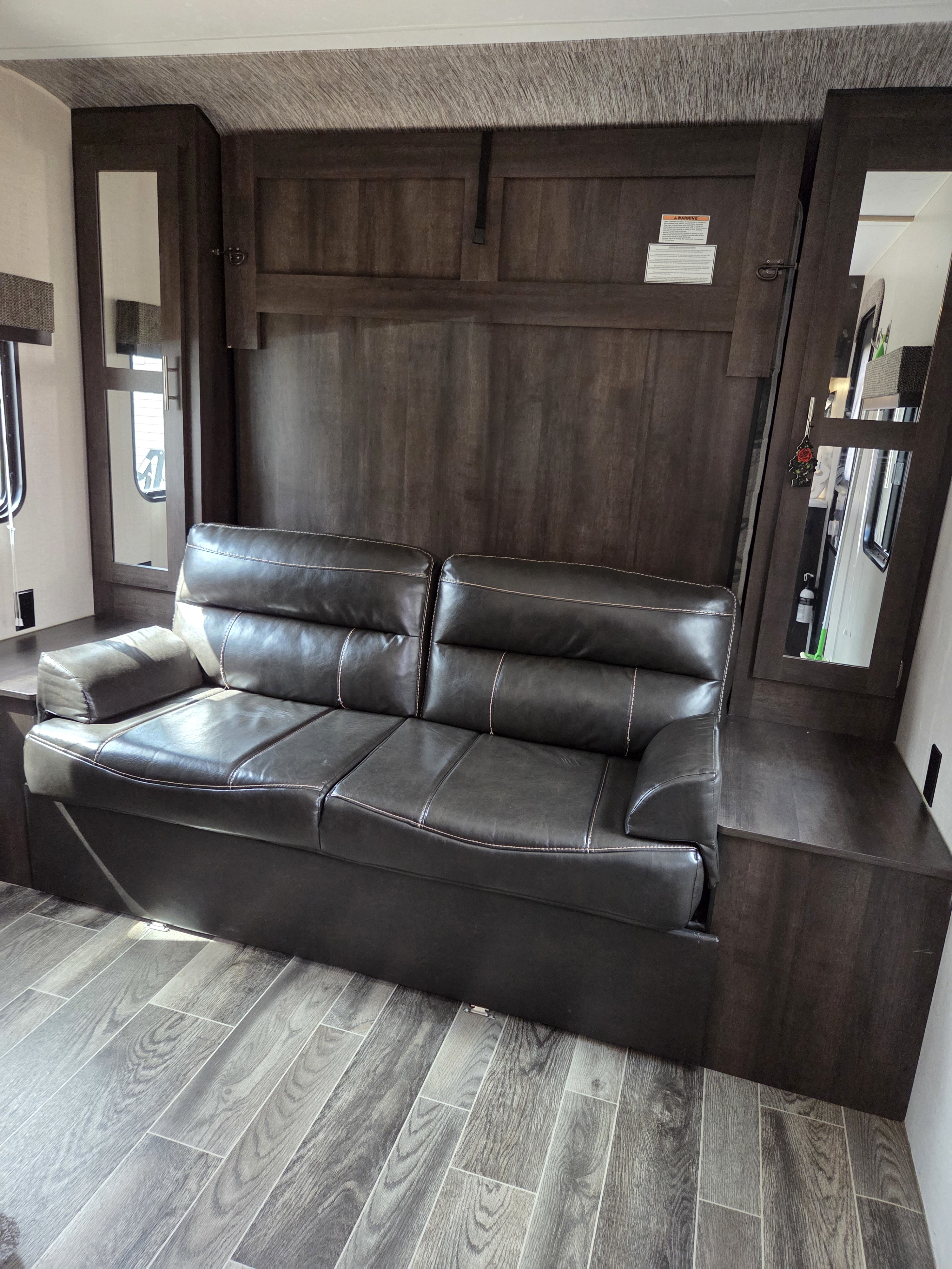 Cozy rv rental interior: dark brown leather sofa against wood walls, flanked by mirrored cabinets, on wood-look flooring.. Sportsmen LE 2018