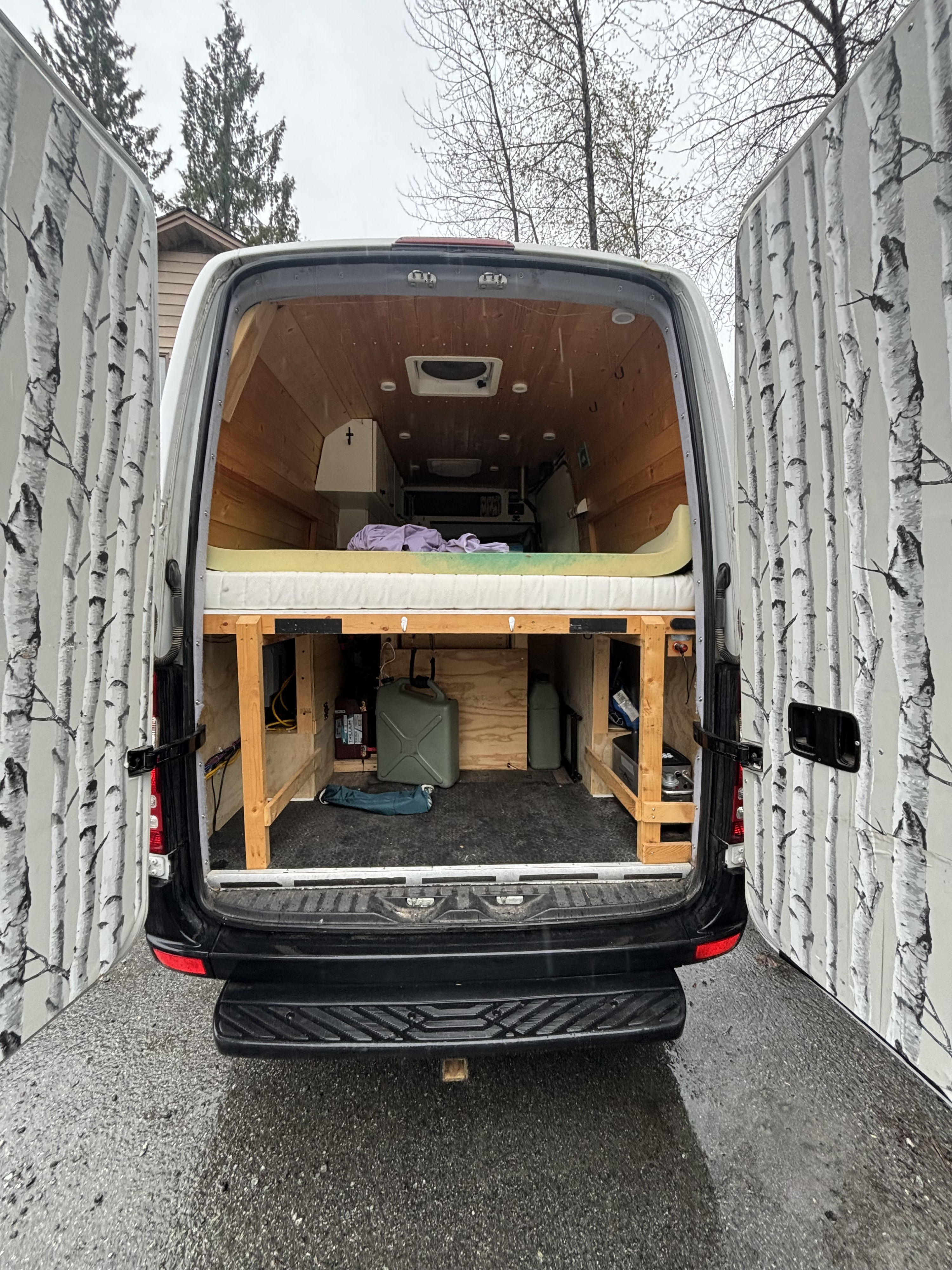 RV rental van's open rear reveals a cozy wooden interior, elevated bed, and storage area. Birch tree doors add flair.. Mercedes-Benz Sprinter 2013
