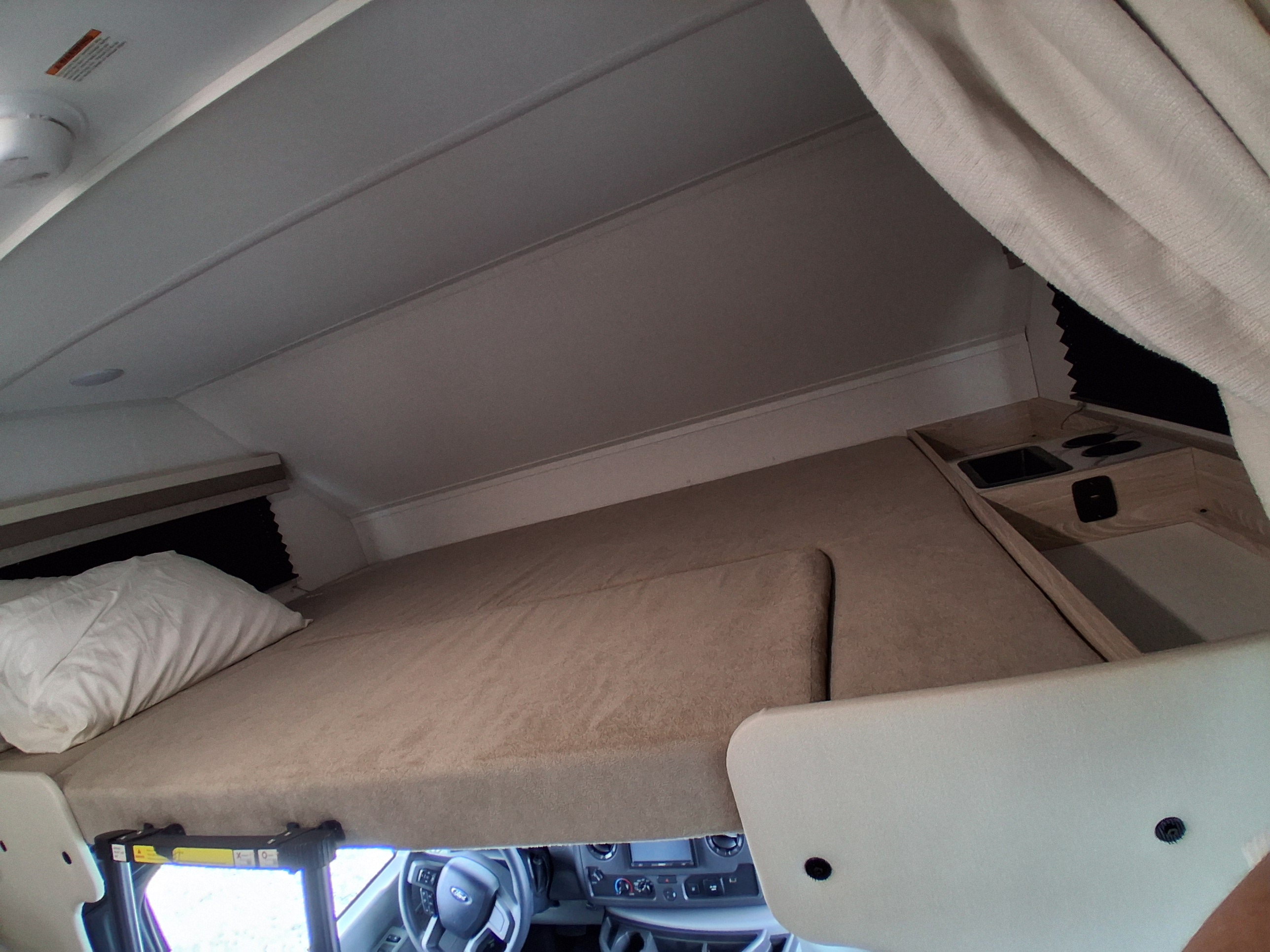Overhead bunk in an rv rental, featuring a pillow, compact sink/stove area, and vehicle dashboard below.. East to West Entrada 2026