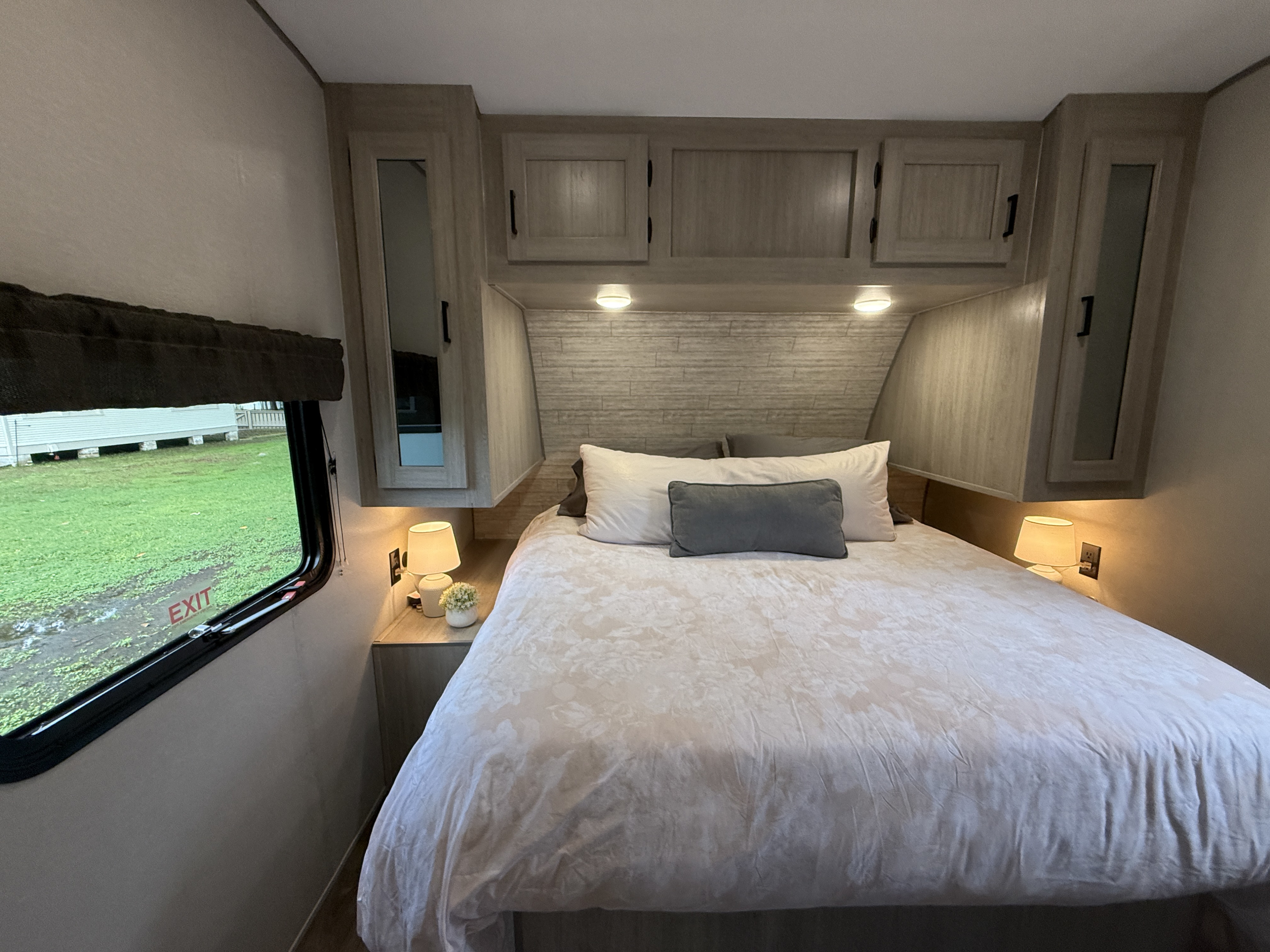 Cozy RV rental bedroom: queen bed with light bedding, modern overhead cabinets, soft bedside lamps, & an exit window.. Coachmen Catalina 2024