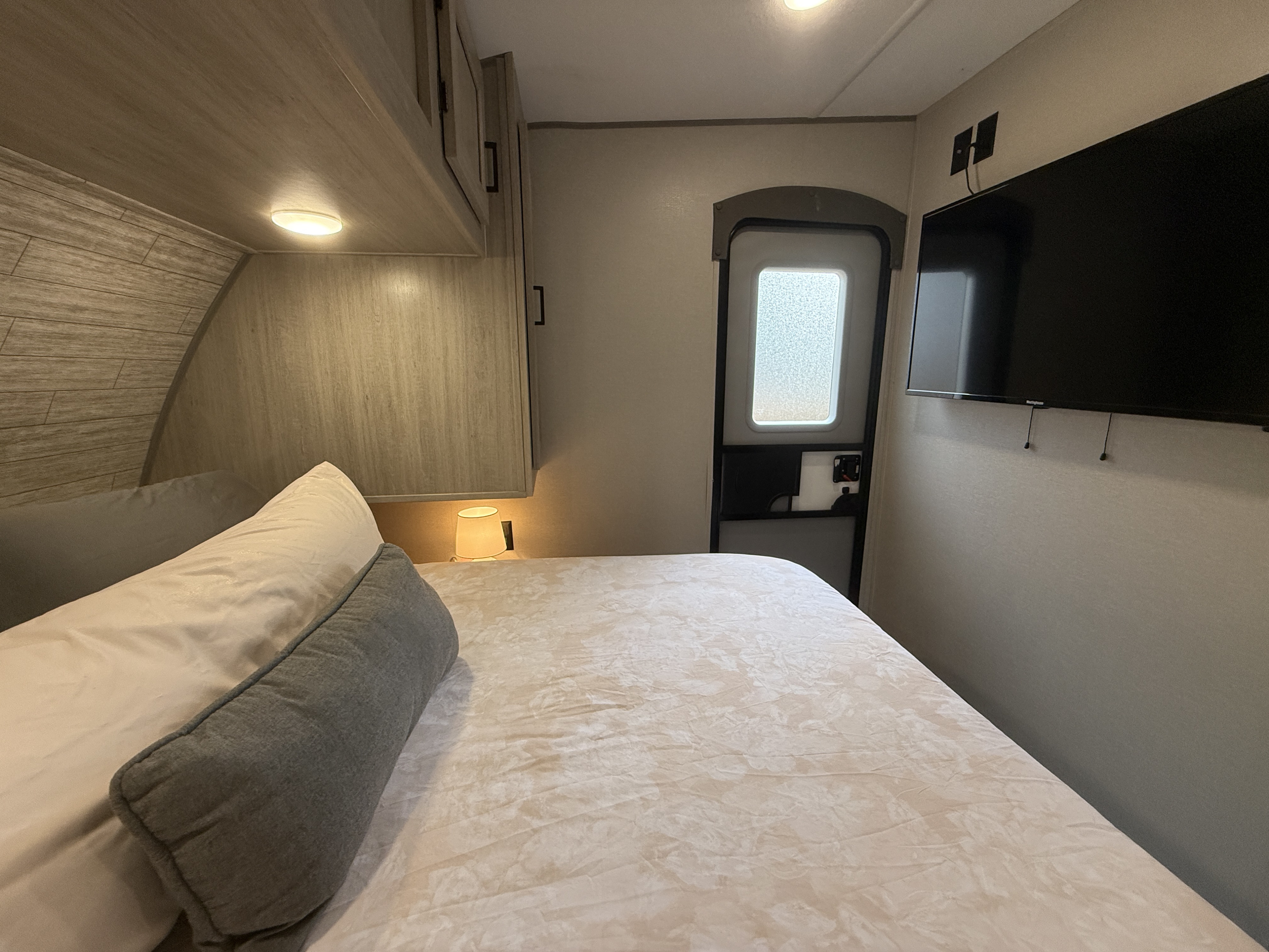 A welcoming RV rental bedroom with a neatly made bed, bedside lamp, wall-mounted TV, overhead storage, and an exit door.. Coachmen Catalina 2024