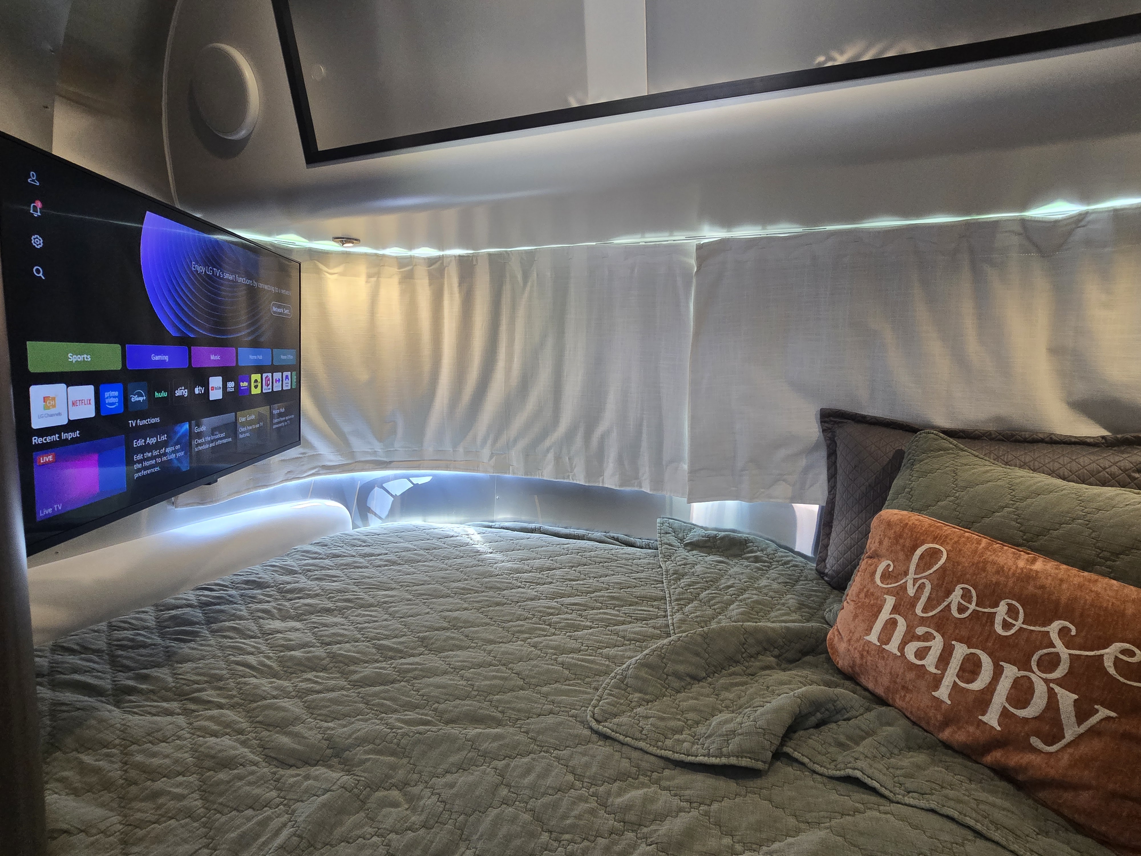 Cozy RV rental bedroom with a smart TV, quilted bed, "choose happy" pillow, and ambient lighting above privacy curtains.. Airstream Caraval 2021