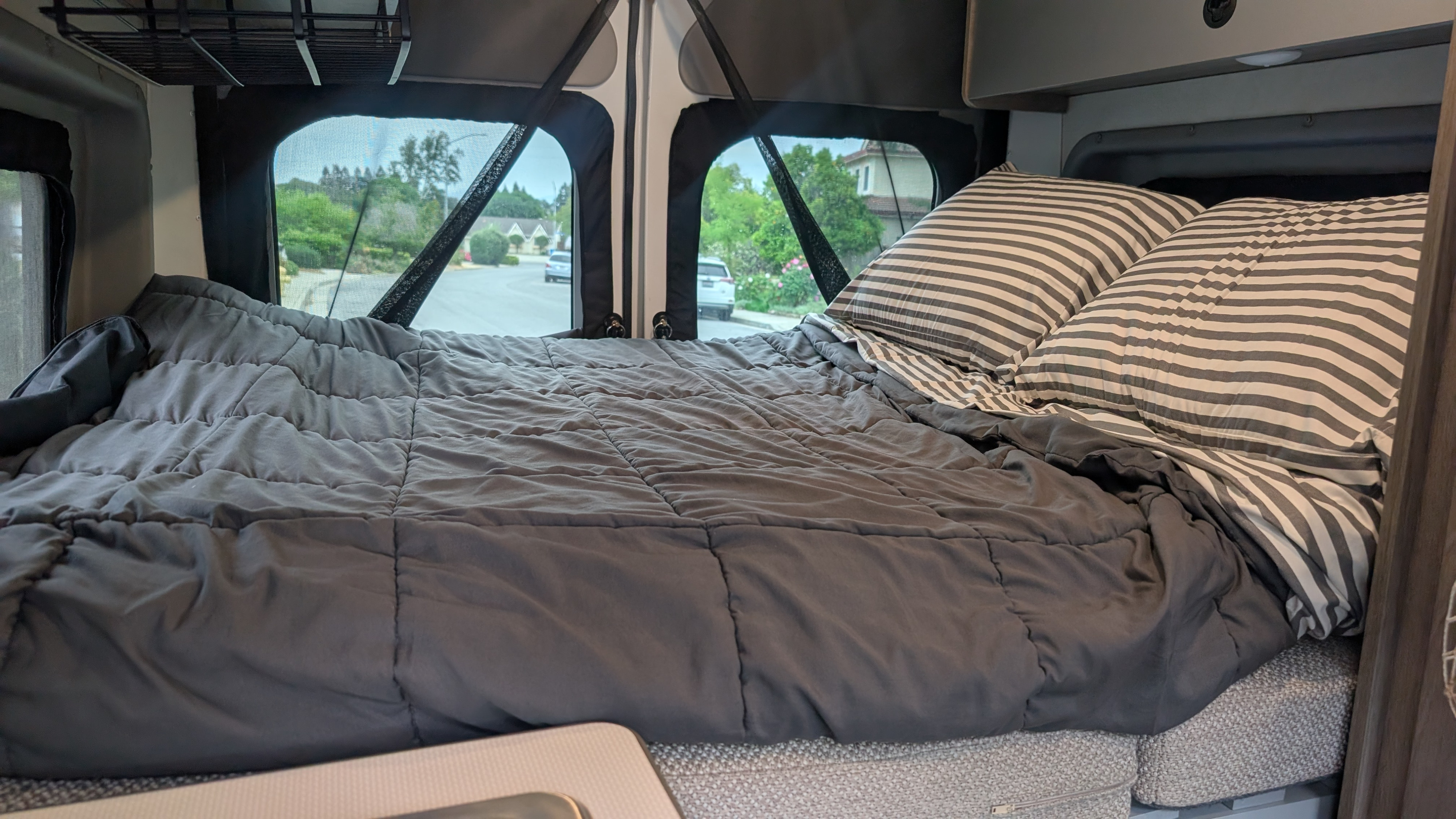 Cozy RV rental bed with grey comforter and striped pillows, offering a view of a residential street outside. Ideal for travel.. Winnebago Solis 2021