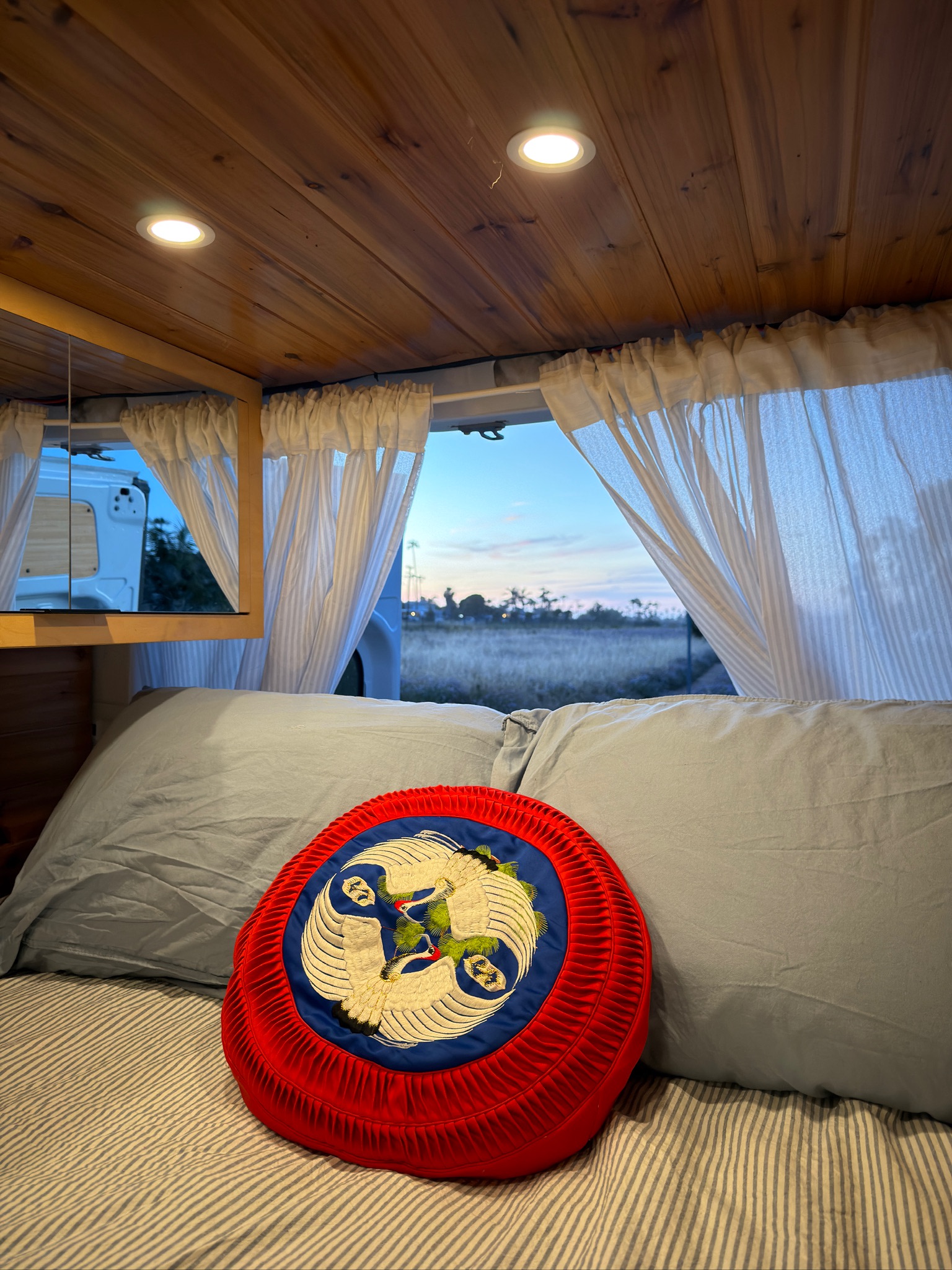 Cozy RV rental bedroom with wooden ceiling, soft lights, and a tranquil view of a field at dusk. Features a unique red embroidered cushion.. Mercedes-Benz Sprinter 2019