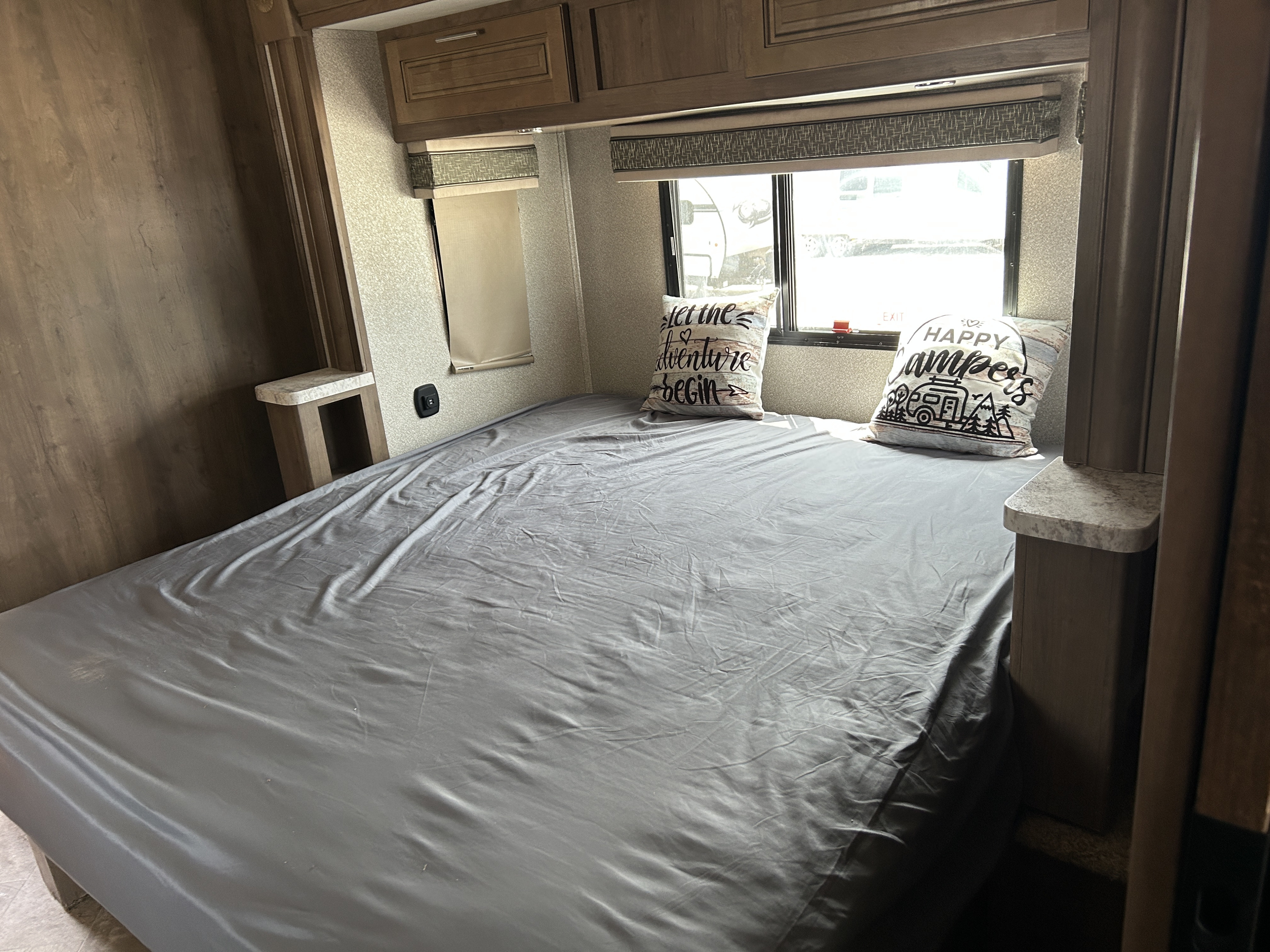 A welcoming rv rental bedroom with a gray bed, "adventure" pillows, wood-toned cabinets, and a window with blinds.. Forest River Georgetown 2019