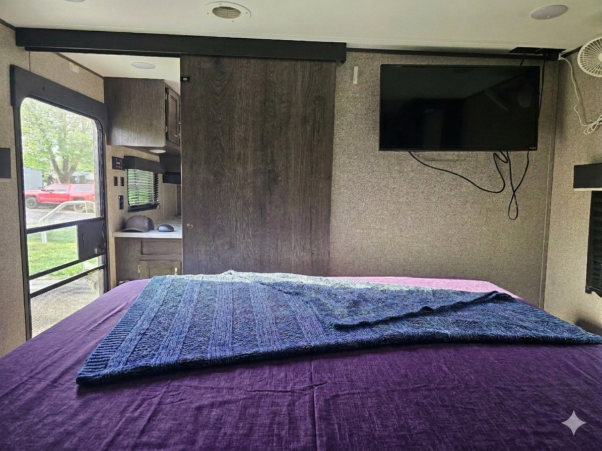 RV rental bedroom interior: purple bed, textured blanket, mounted TV. An open door reveals another RV section and outdoors.. Jayco Jay Flight SLX 2020