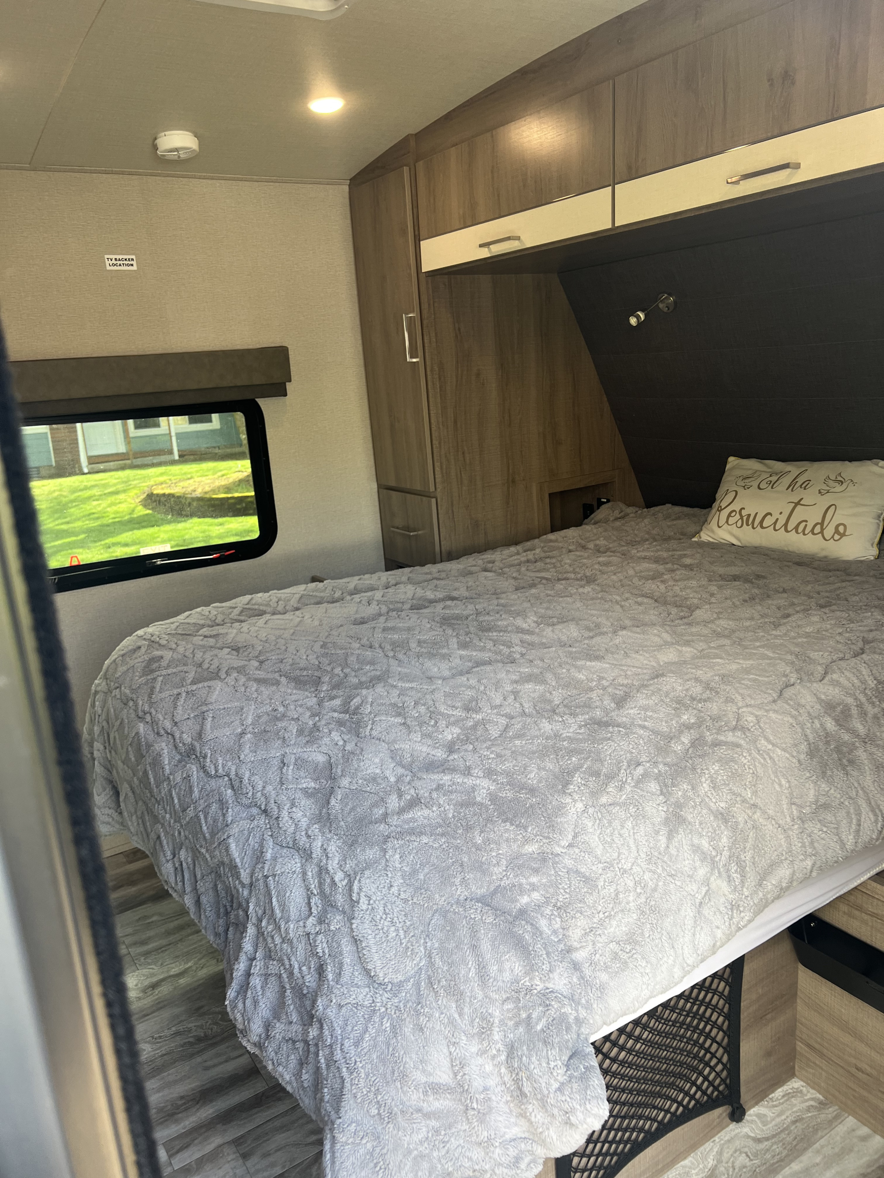 Cozy RV rental bedroom featuring a plush bed with a grey textured comforter, overhead cabinets, and a window showing greenery.. Grand Design Imagine 2022
