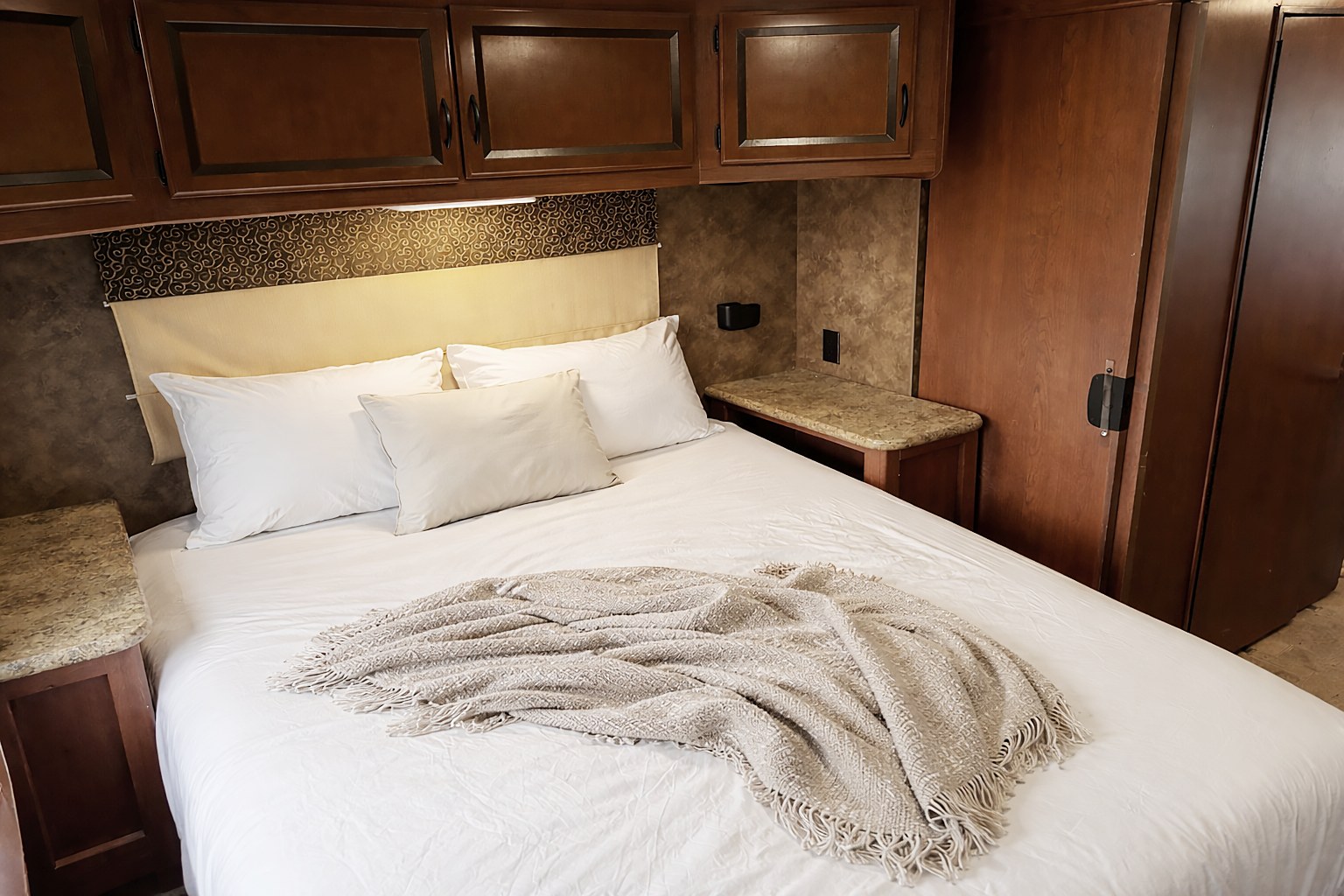 An RV rental bedroom features a large bed with white linens, flanked by small counters. Dark wood overhead cabinets provide storage.. Thor Hurricane 2014