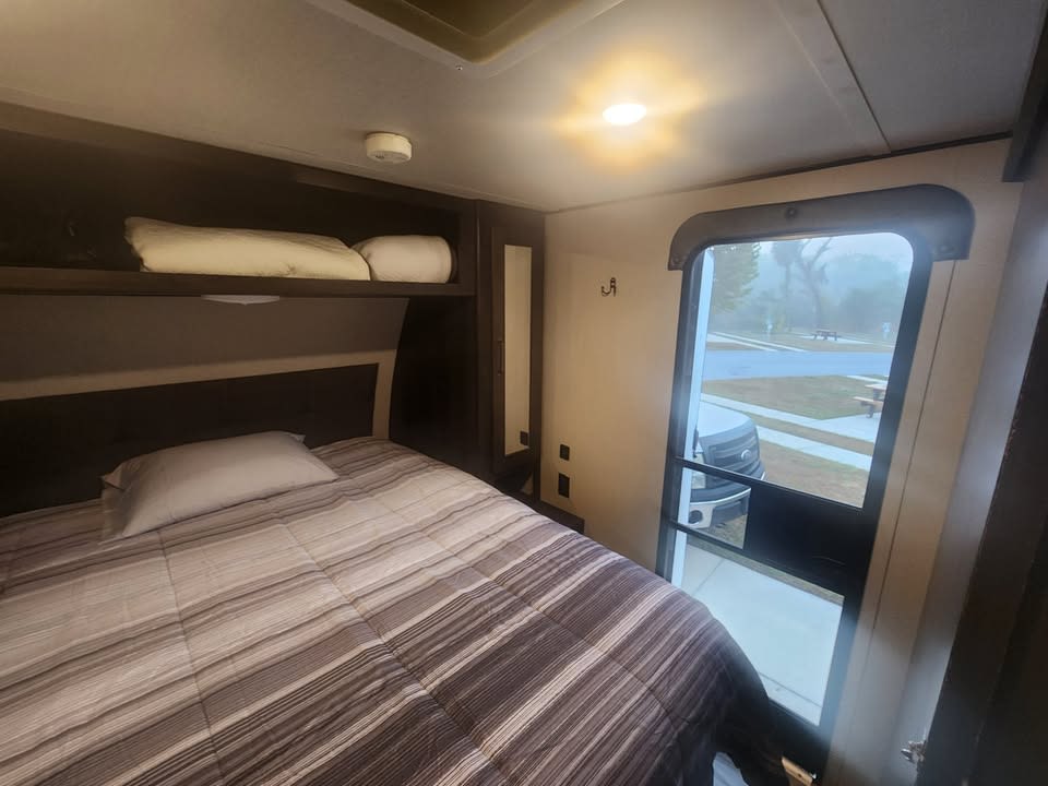 RV rental bedroom with bed, overhead shelf, and a door showing a foggy outdoor view. Compact and ready for travel.. Grand Design Transcend Xplor 2021
