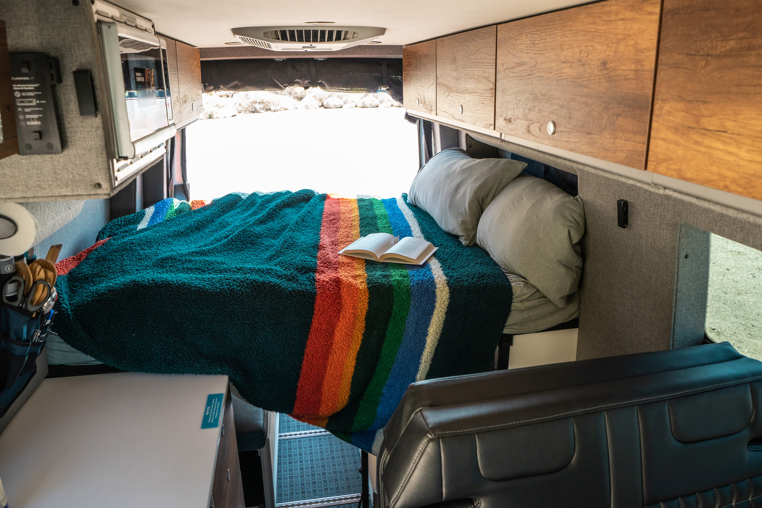 Inside an RV rental: A comfy bed with a rainbow-striped blanket, pillows, and a book. Open back doors show a bright outdoor scene.. Storyteller Overland Storyteller Overland Class B 2022