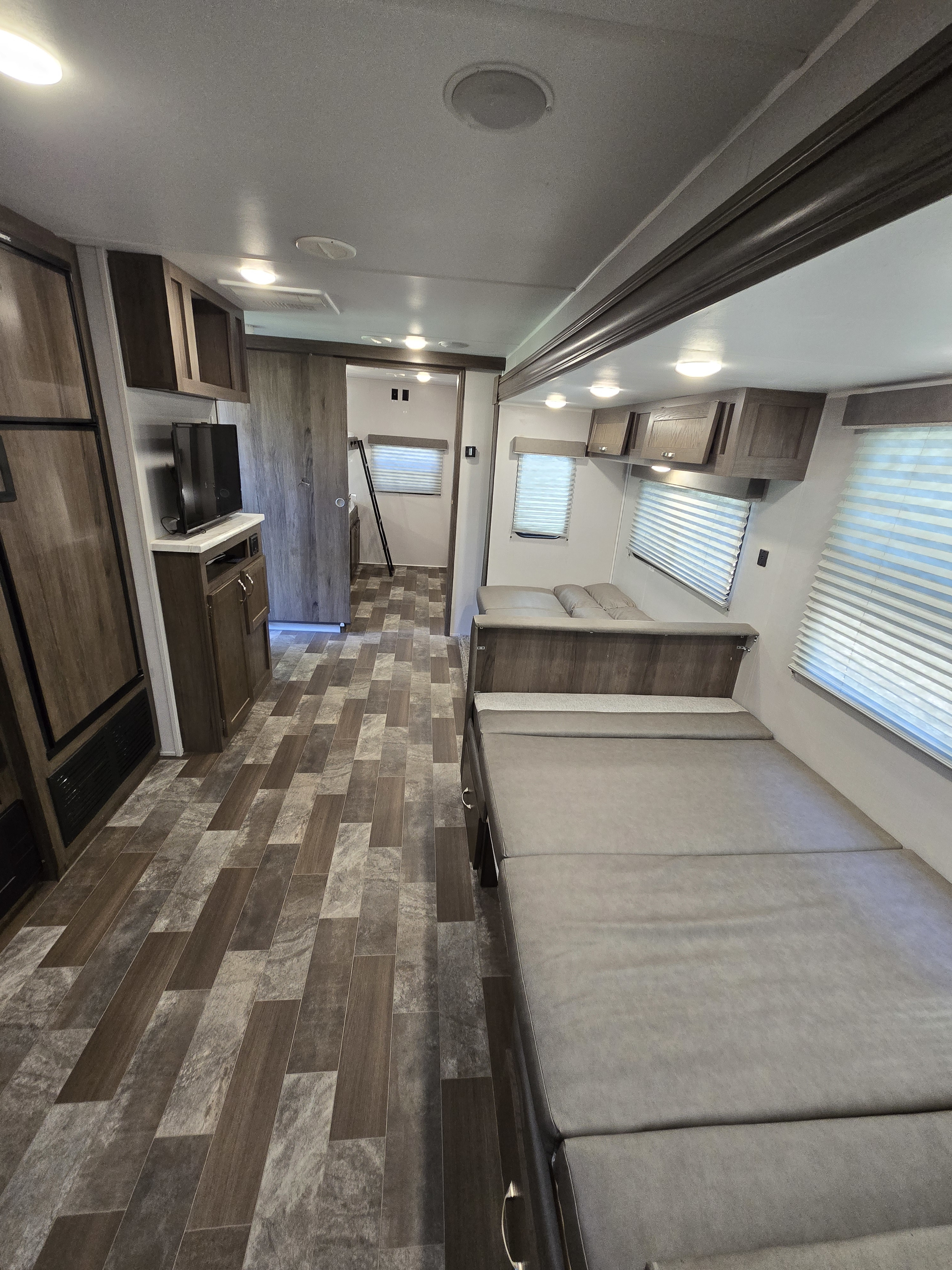 RV rental interior: Main living/sleeping area with gray seating/bed, wood-look cabinets, TV, and rear bedroom beyond. Patterned flooring.. Forest River Silver Lake East-West 2020