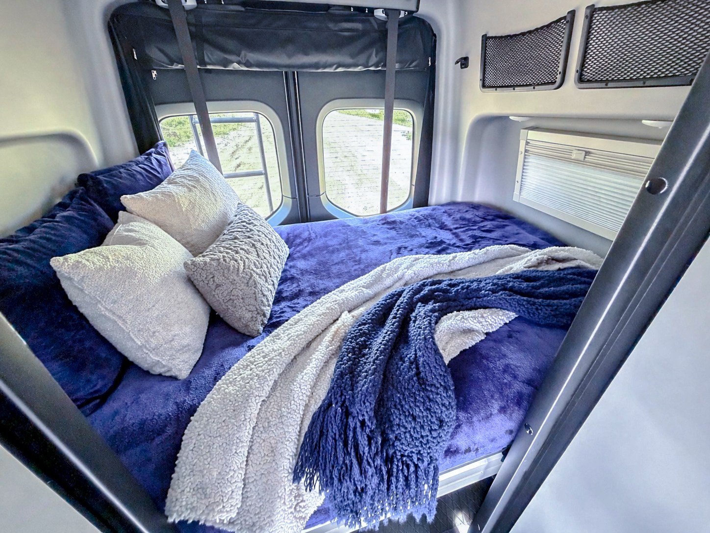 This cozy rv rental bed features plush purple bedding, white and gray textured pillows, and warm blankets. Ideal for travel.. Winnebago Revel 2026