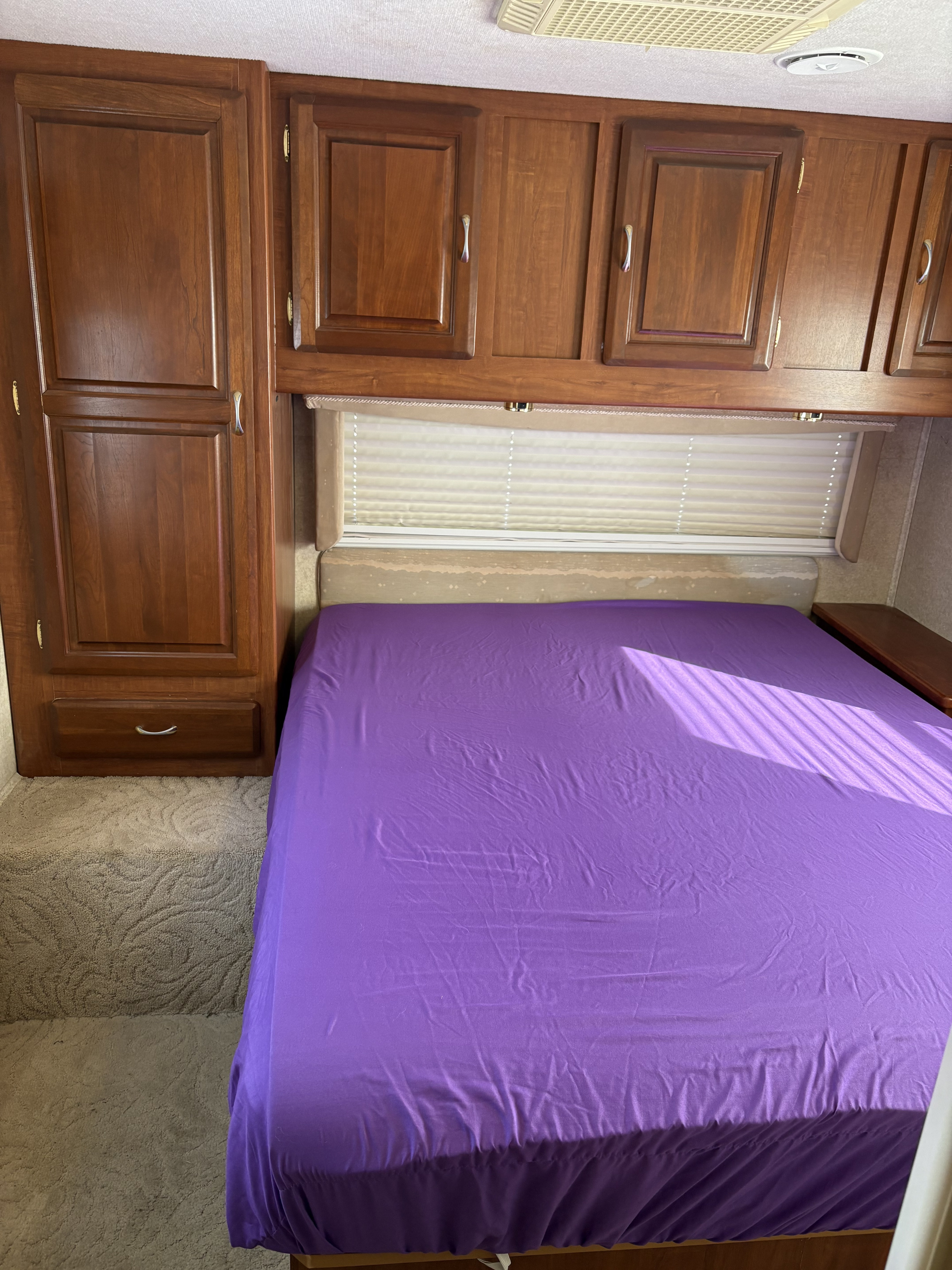 RV rental bedroom with a bed in bright purple sheets, overhead wooden cabinets, and a tall wardrobe. Sunlight falls across the bed.. Jayco Granite Ridge 2004