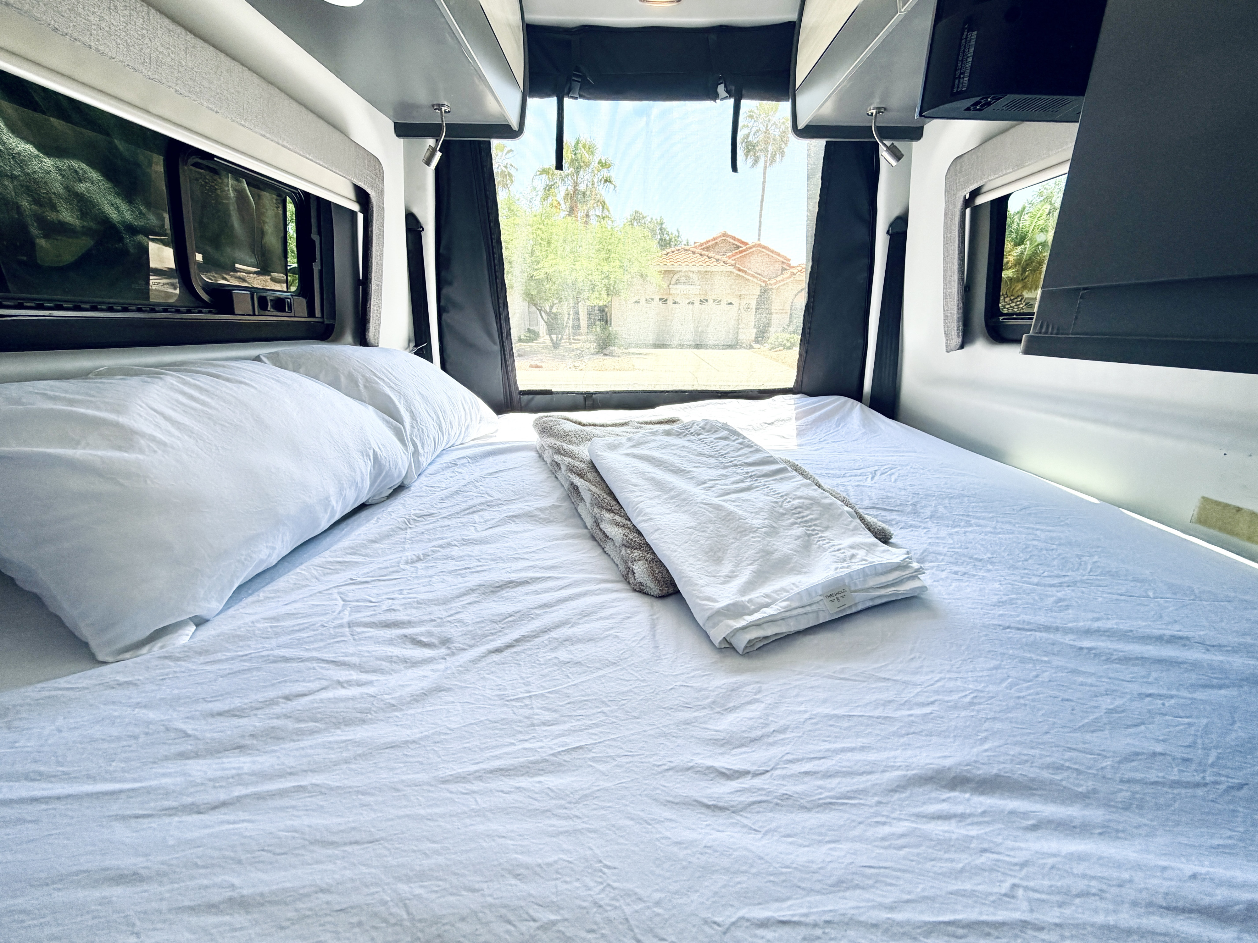 Cozy rv rental bed, dressed in white sheets & pillows, with a comfy throw. The back window reveals a sunny suburban street.. Thor Synergy 2022