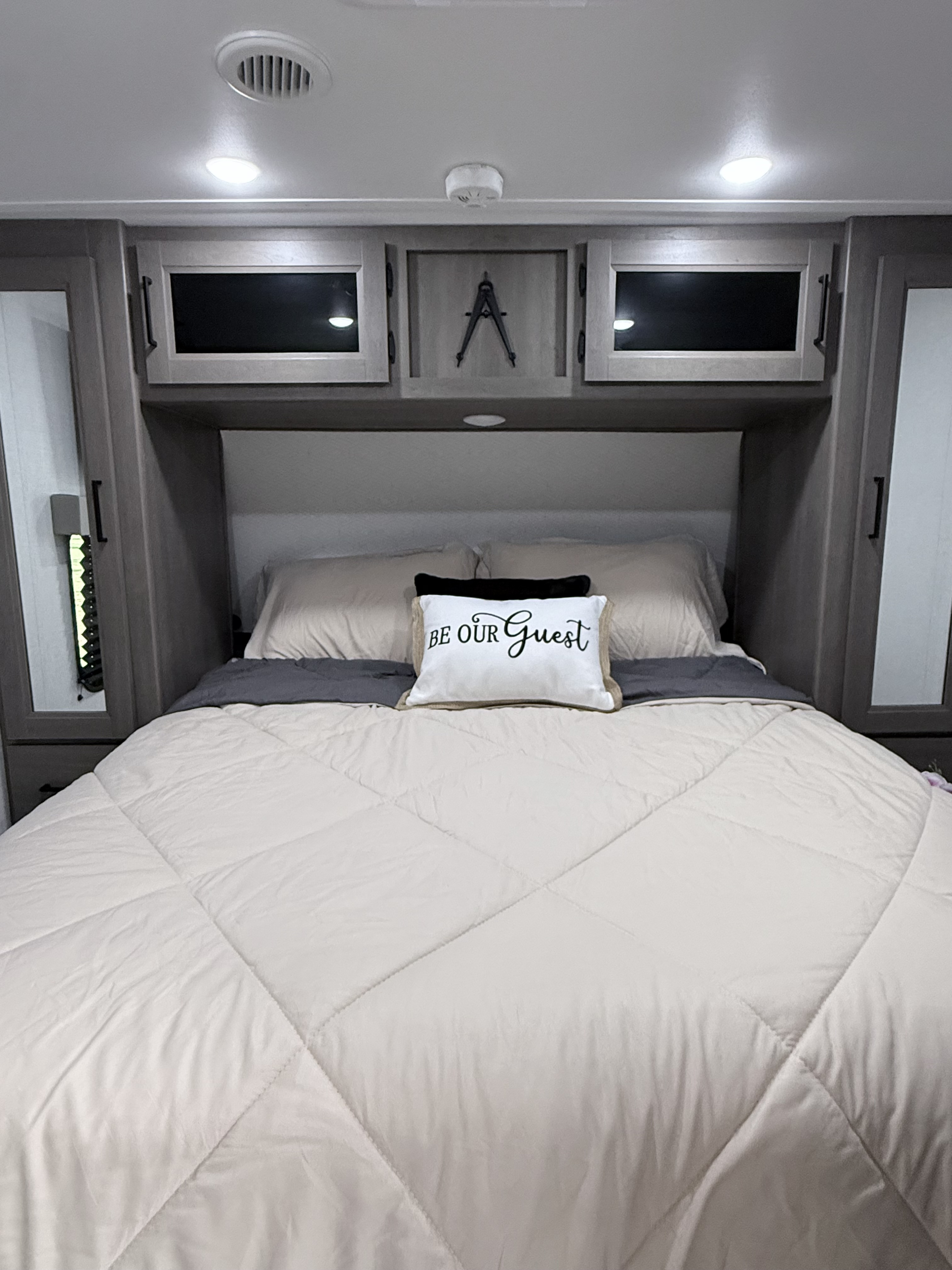 Neat RV rental bedroom interior, showcasing a comfortable bed with a "Be Our Guest" pillow and practical overhead cabinets.. Grand Design Transcend 2025
