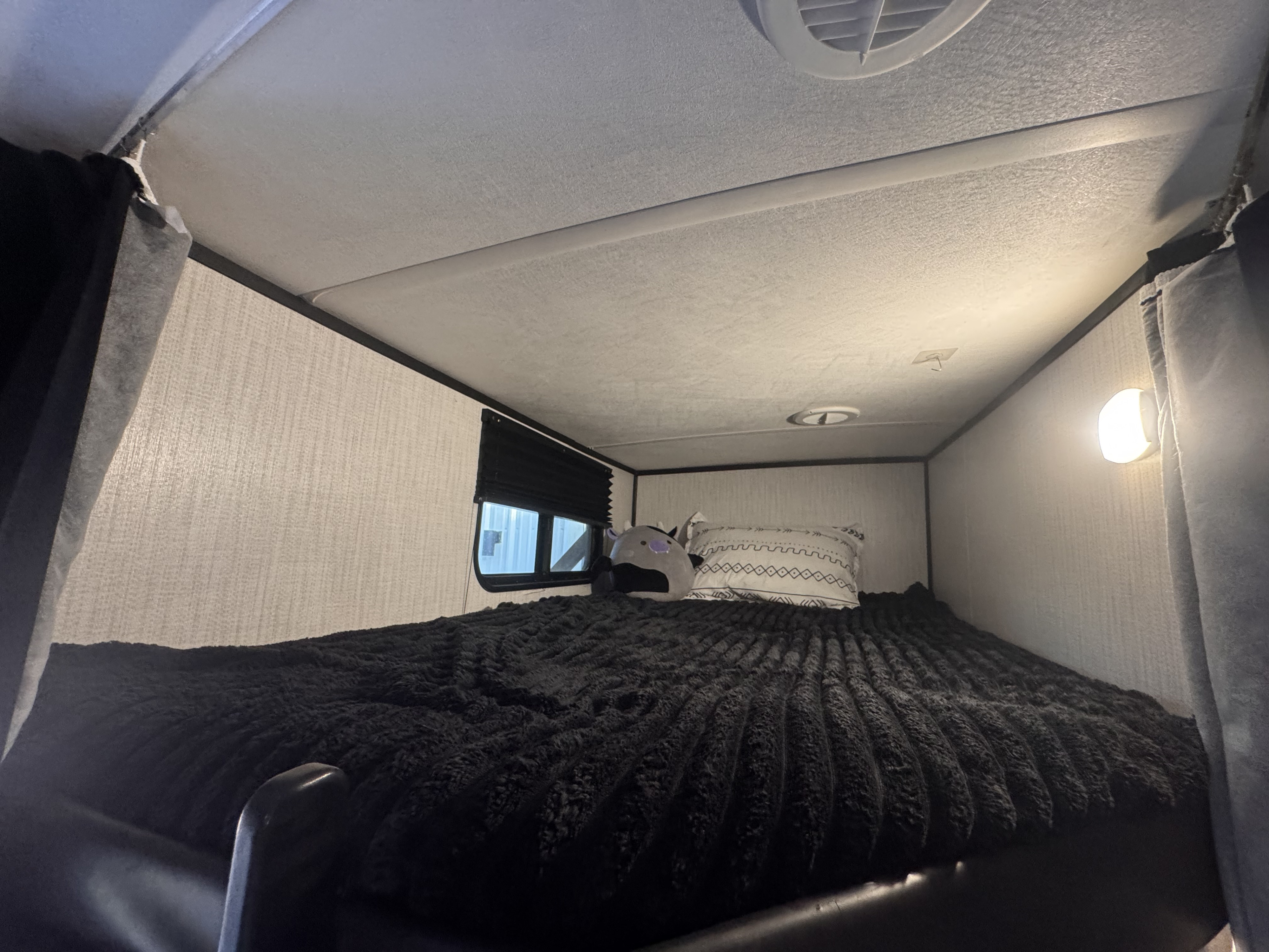 Cozy RV rental bunk: a sleeping area with a plush black blanket, patterned pillow, window, and wall light for travel comfort.. Heartland RVs North Trail 2021