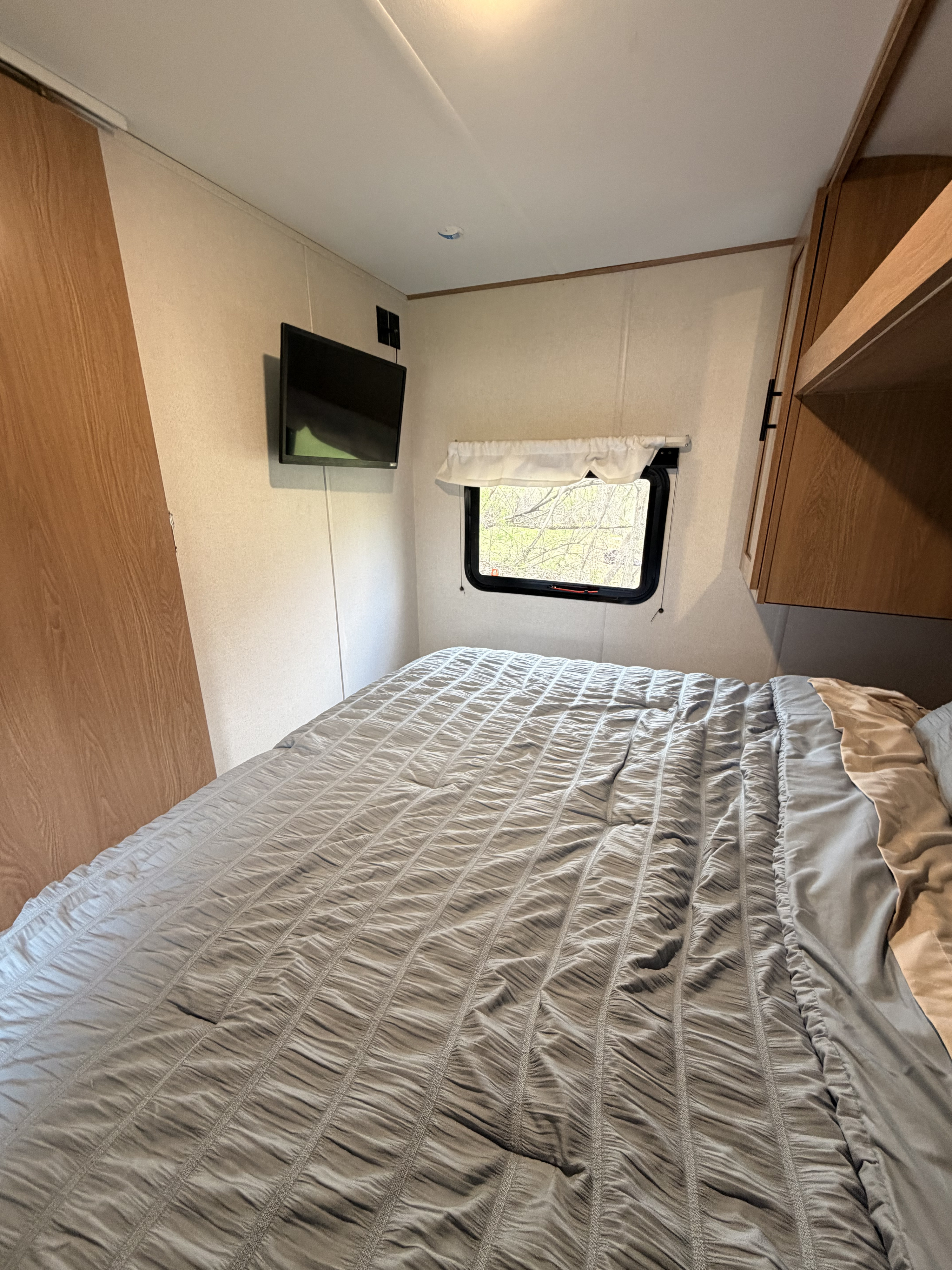 This cozy RV rental bedroom features a comfy bed with a gray comforter, a window looking out to trees, a mounted TV, and overhead cabinets.. Avenger LE 2022
