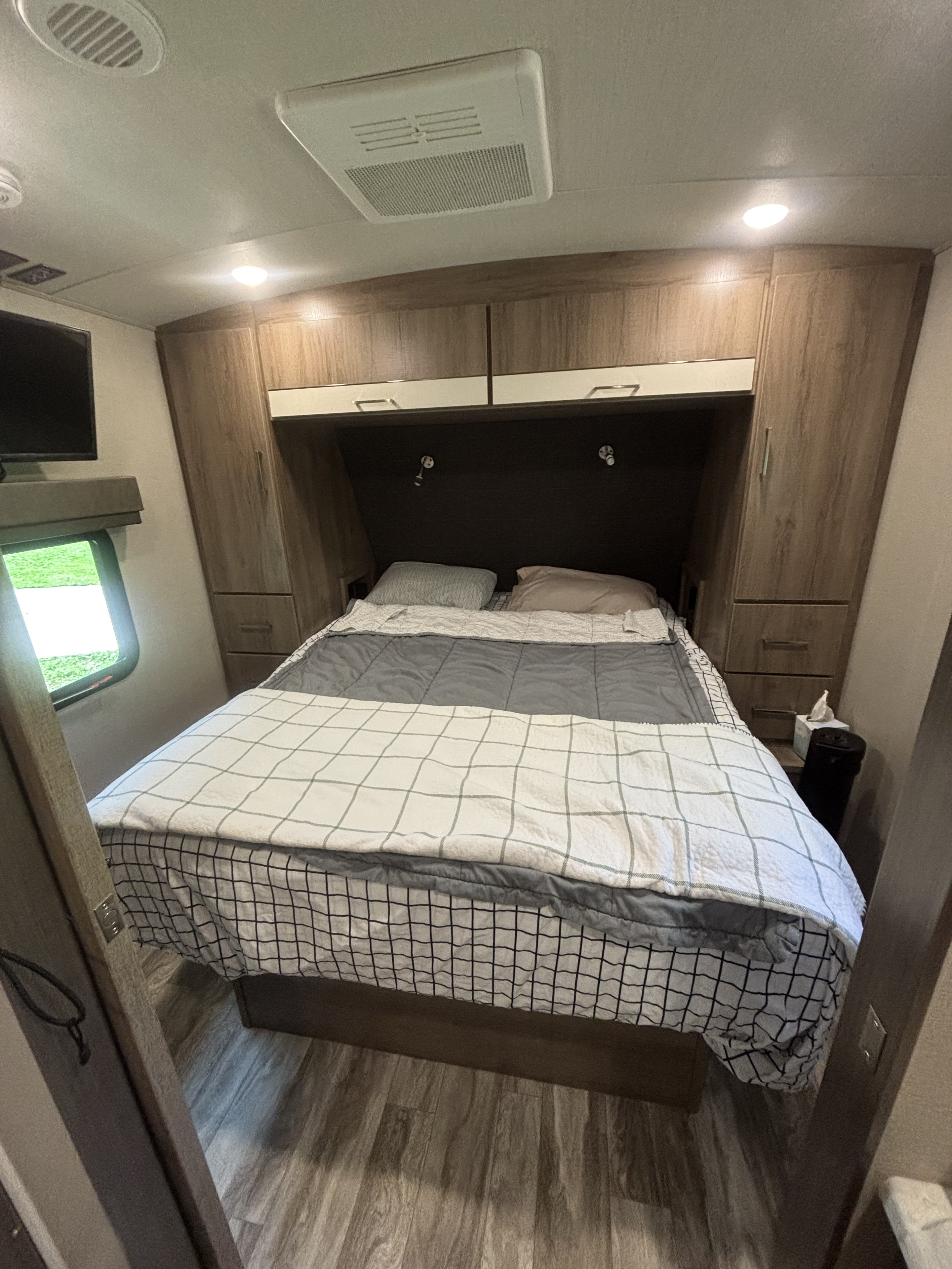 RV rental bedroom: cozy interior with bed, overhead/side cabinets, reading lights, window, TV, and wood-look flooring.. Grand Design Imagine 2023