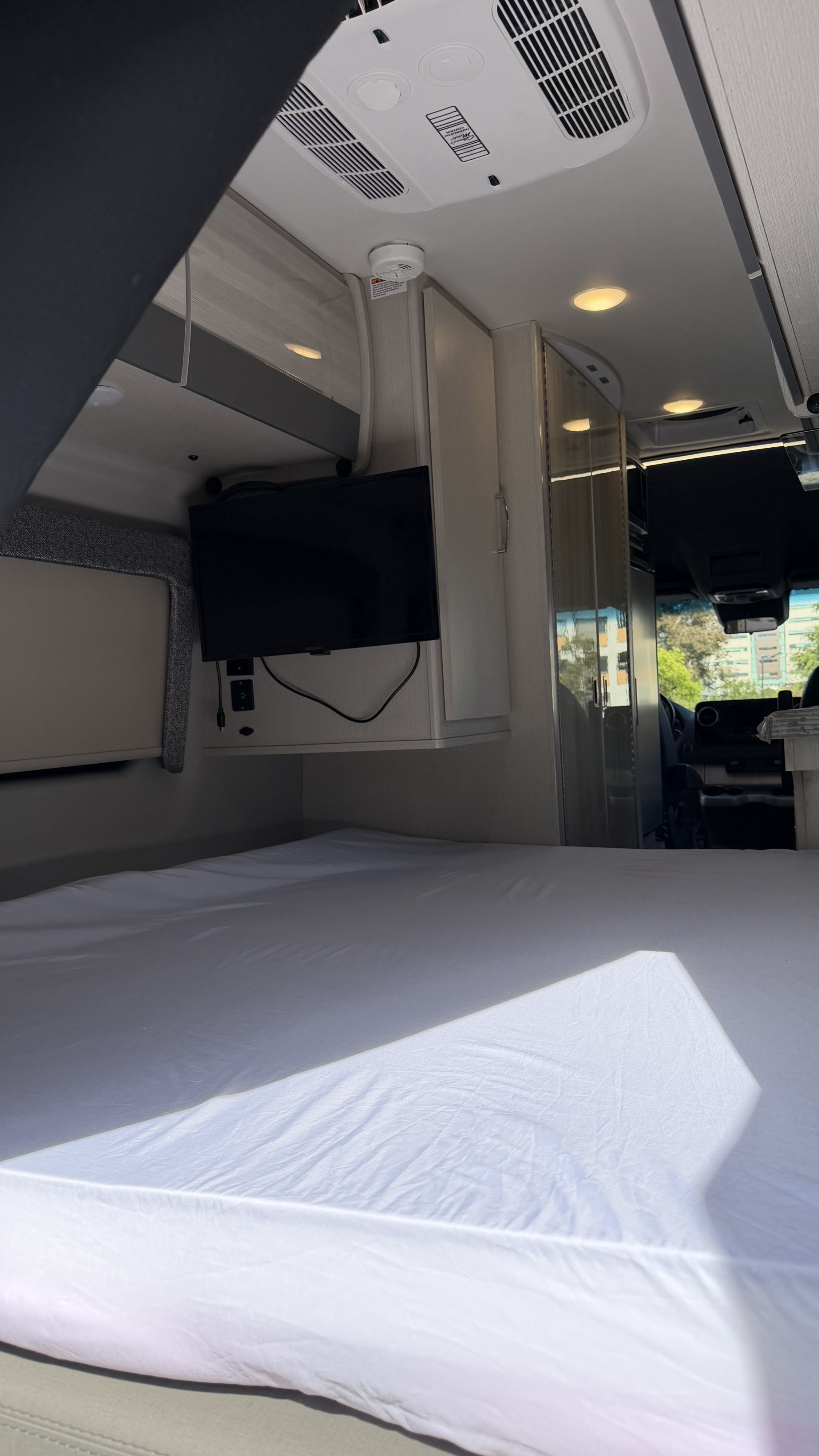 Relax in this rv rental! Modern camper van interior with a bed, mounted TV, and overhead AC unit. Sunlit sleeping area.. Thor Tranquility 2023
