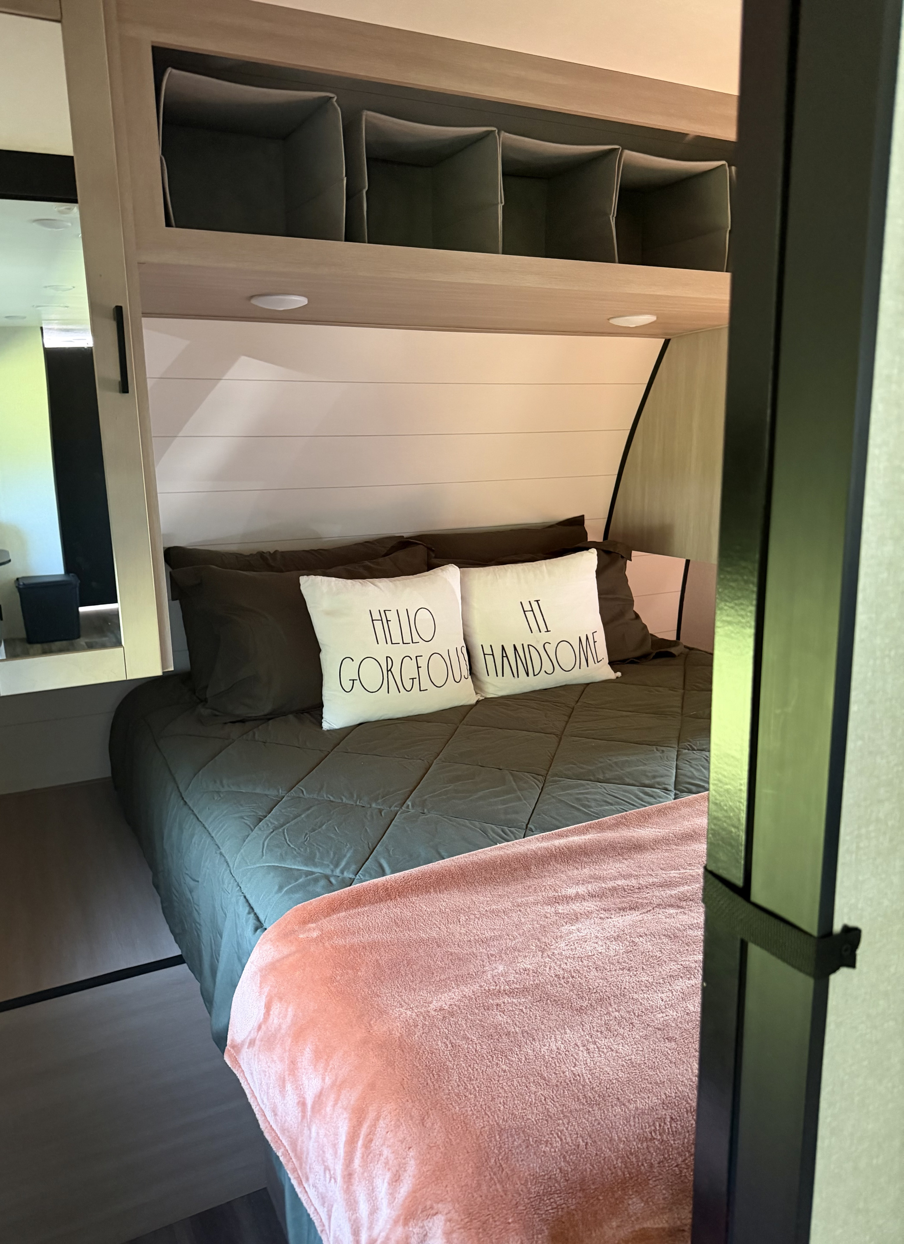 Cozy RV rental bedroom featuring a dark green bed with "HELLO GORGEOUS" pillows, a pink throw, and overhead storage.. Highland Ridge RV Colt 2026