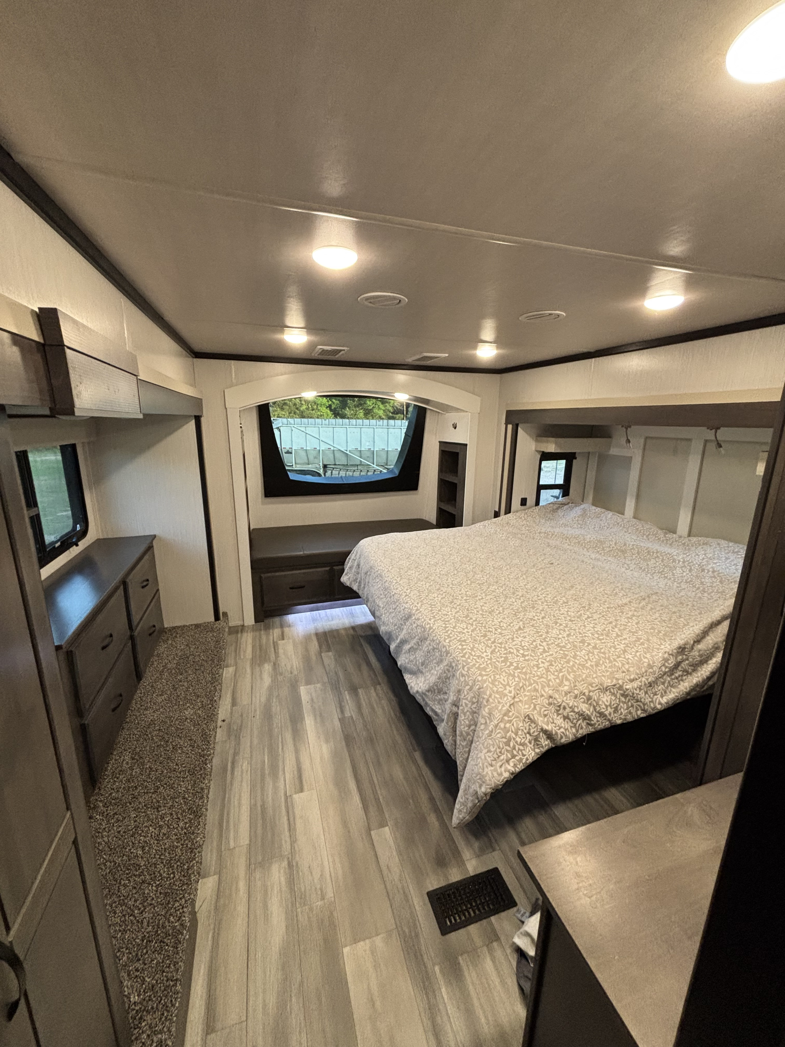 Inside an RV rental: a cozy bedroom with a patterned bed, modern wood-look floors, a dresser, and a panoramic window.. Cedar Creek Champagne 2022