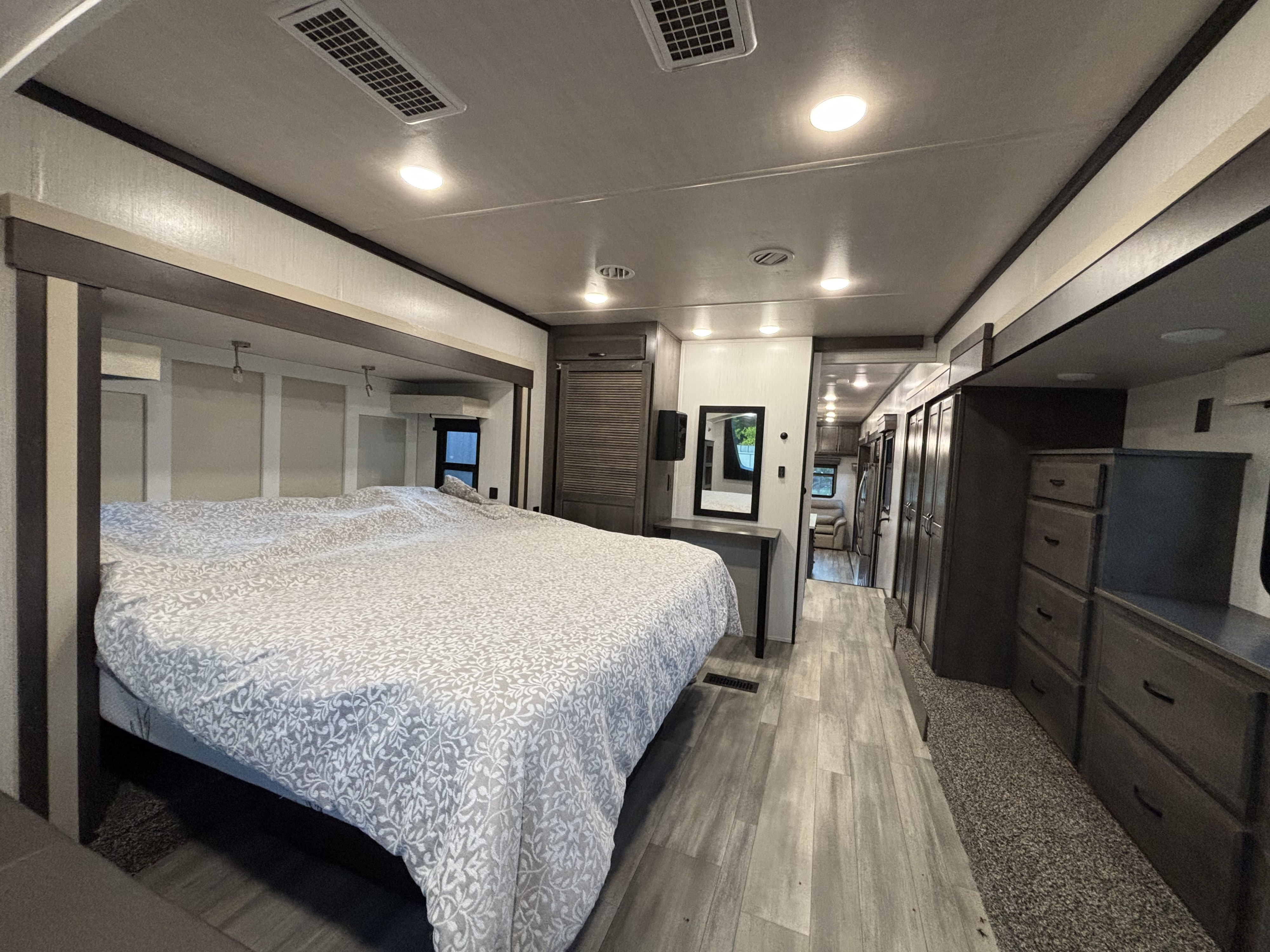 Spacious and modern RV rental bedroom interior with a large bed, dresser, and mirror, leading to a well-lit hallway.. Cedar Creek Champagne 2022