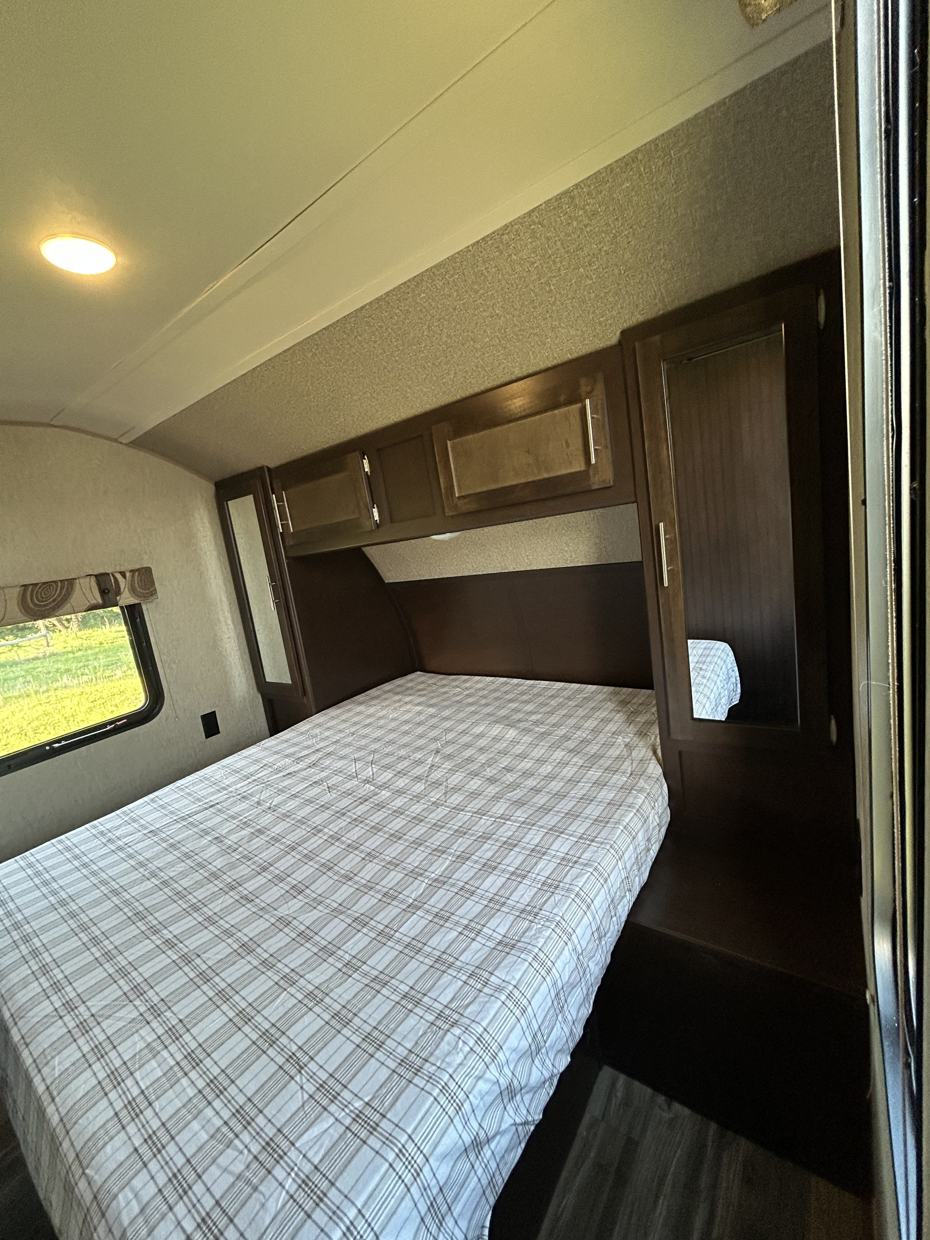 This RV rental bedroom shows a bed with plaid sheets, built-in dark wood cabinets, and a window overlooking greenery.. Forest River Cherokee 2016
