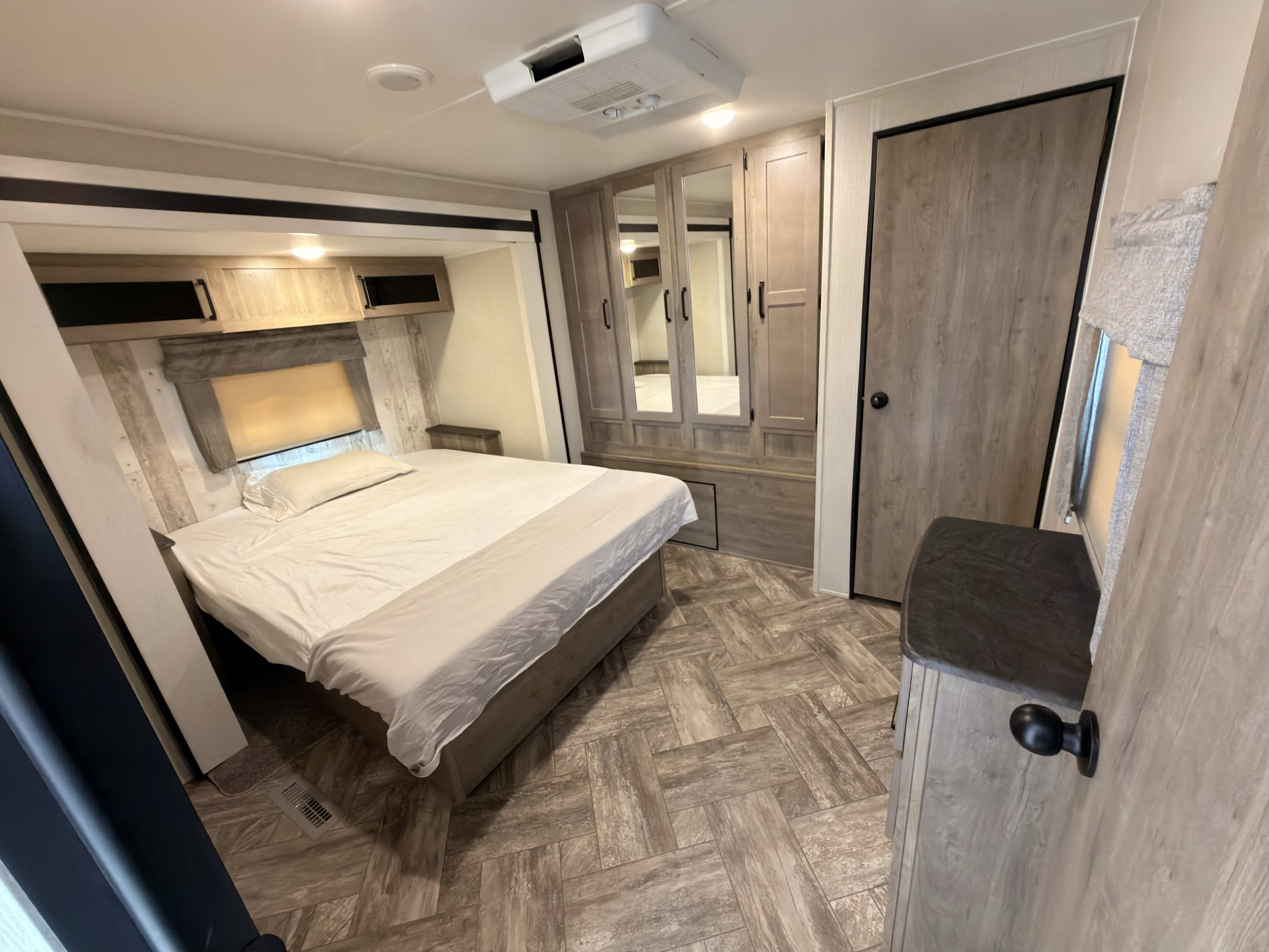 This inviting RV rental bedroom features a comfortable bed, overhead cabinets, a mirrored wardrobe, and patterned wood-look flooring.. Forest River Puma 2023