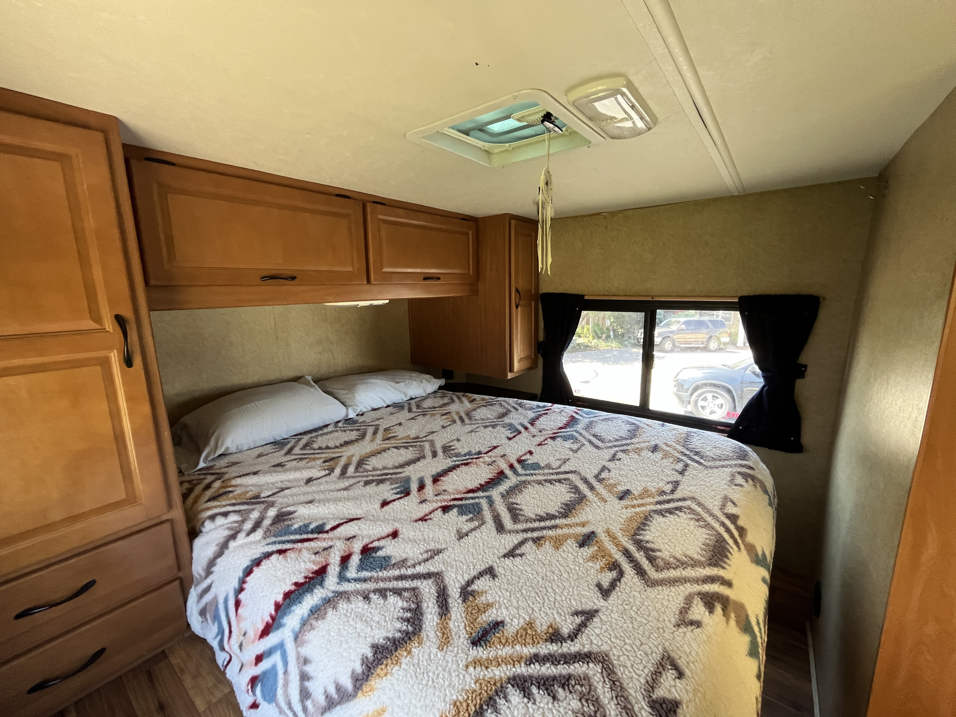 A cozy RV rental bedroom features a comfortable bed with a patterned blanket, overhead storage, and a window with curtains.. Thor Majestic 2016