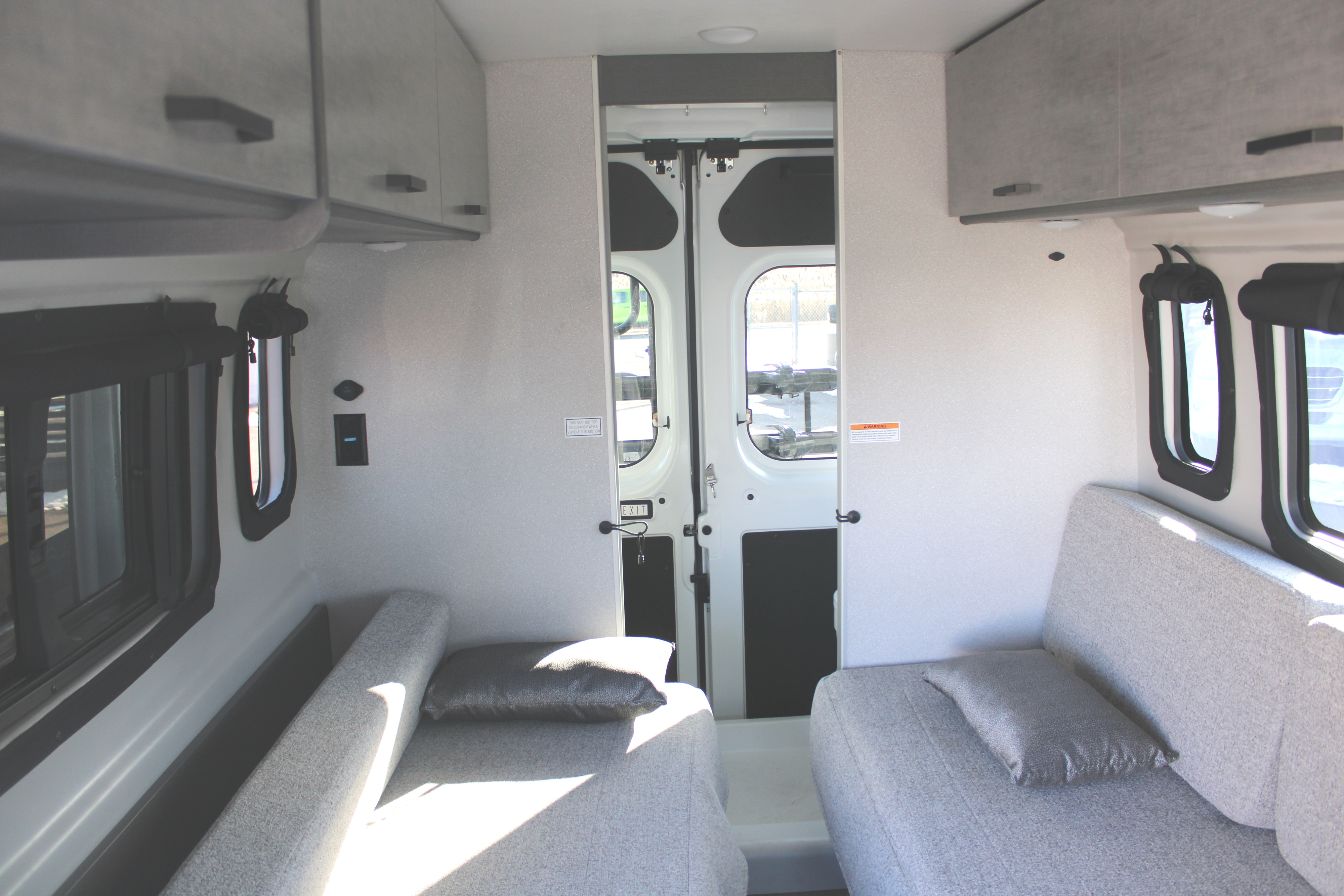 View inside a spacious rv rental camper van, featuring light grey benches, overhead cabinets, and rear access doors.. Thor Motor Coach Tellaro 2026