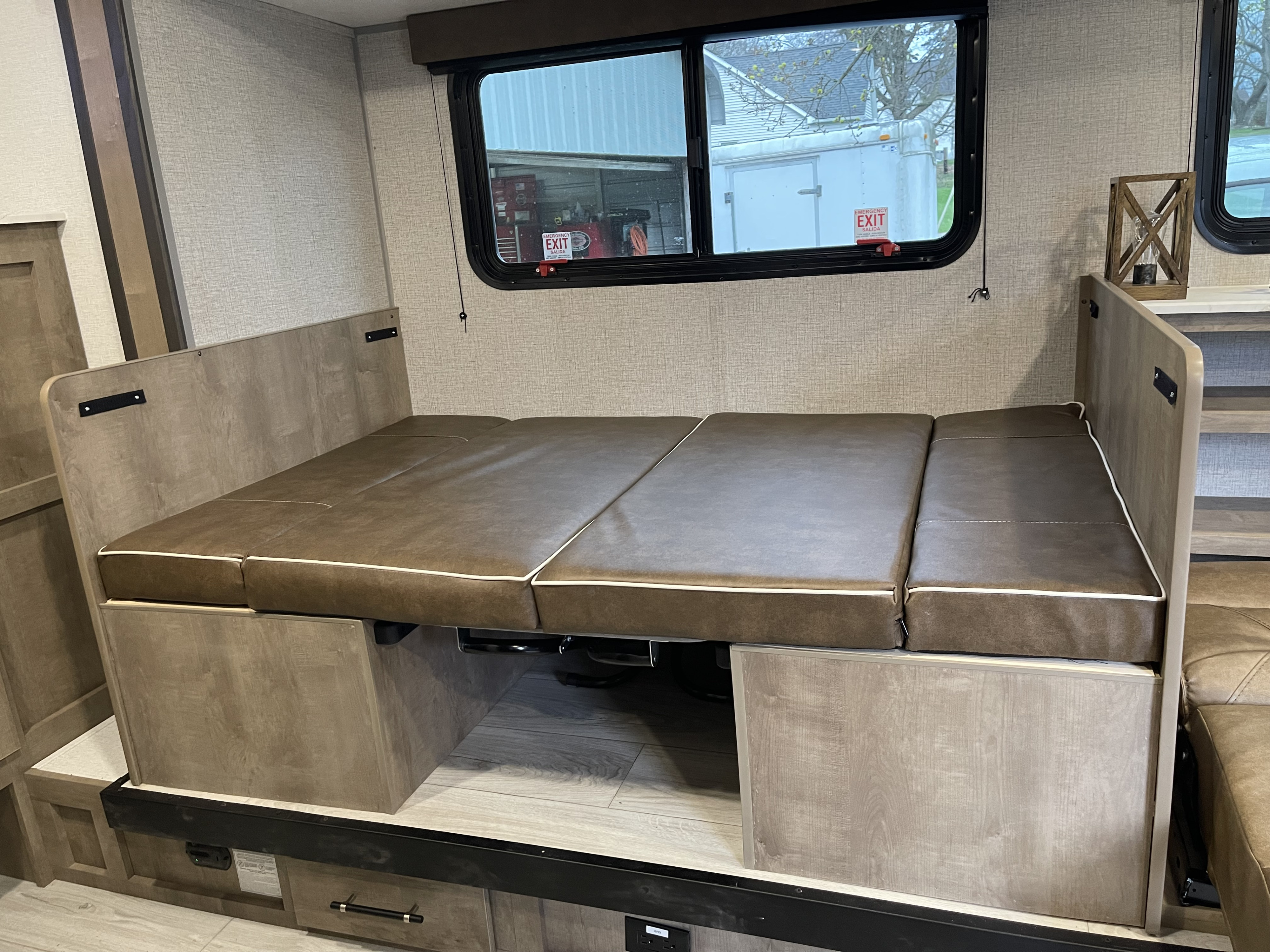 RV rental interior shows a dinette folded into a bed with brown cushions and light wood finishes, beside an emergency exit window.. Keystone RV Springdale Classic 2025