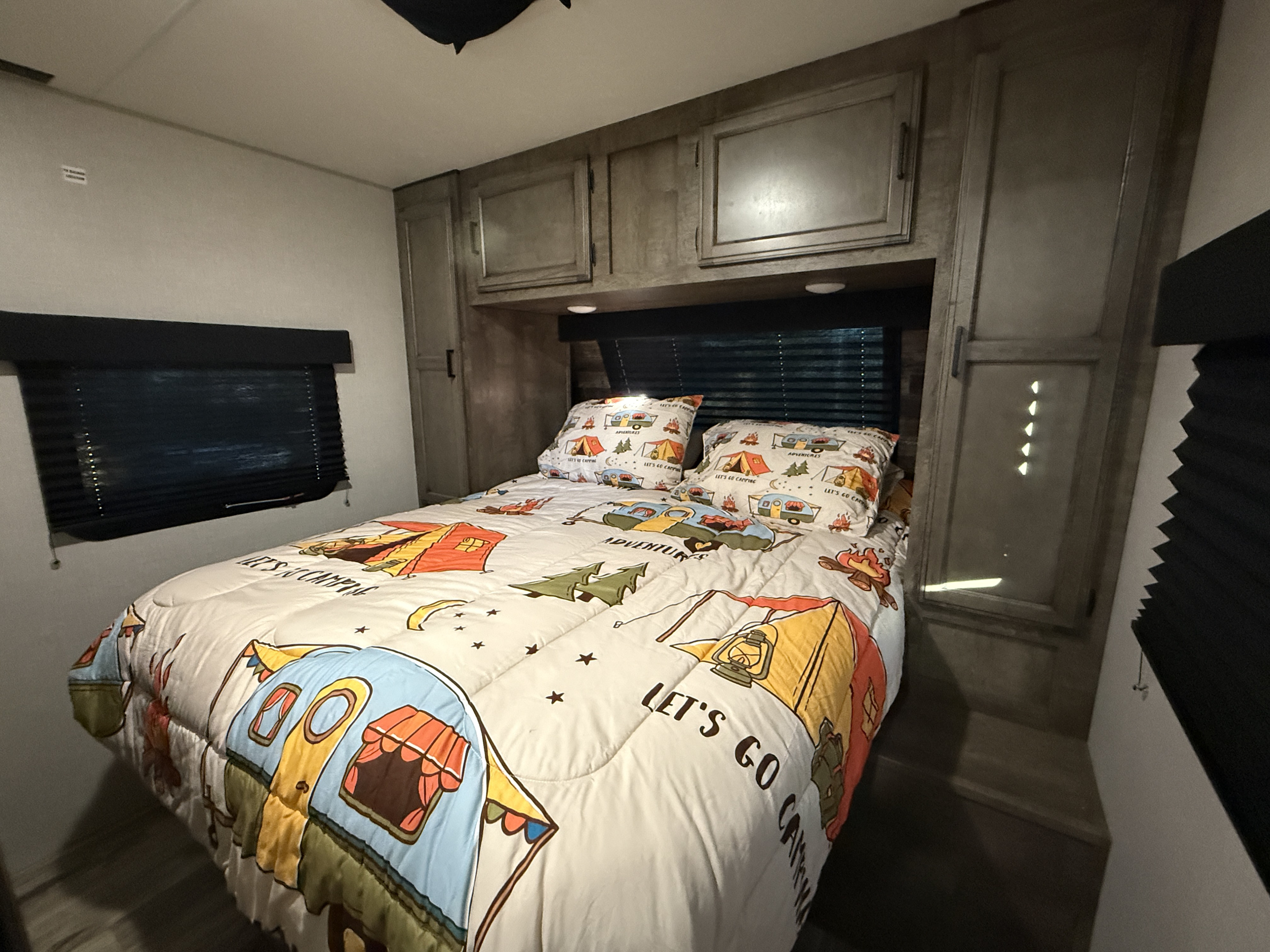 Cozy RV rental bedroom featuring a camping-themed comforter and pillows, overhead cabinets, and window blinds. Ready for adventures!. Sport Trek Sport Trek Trailer 2021