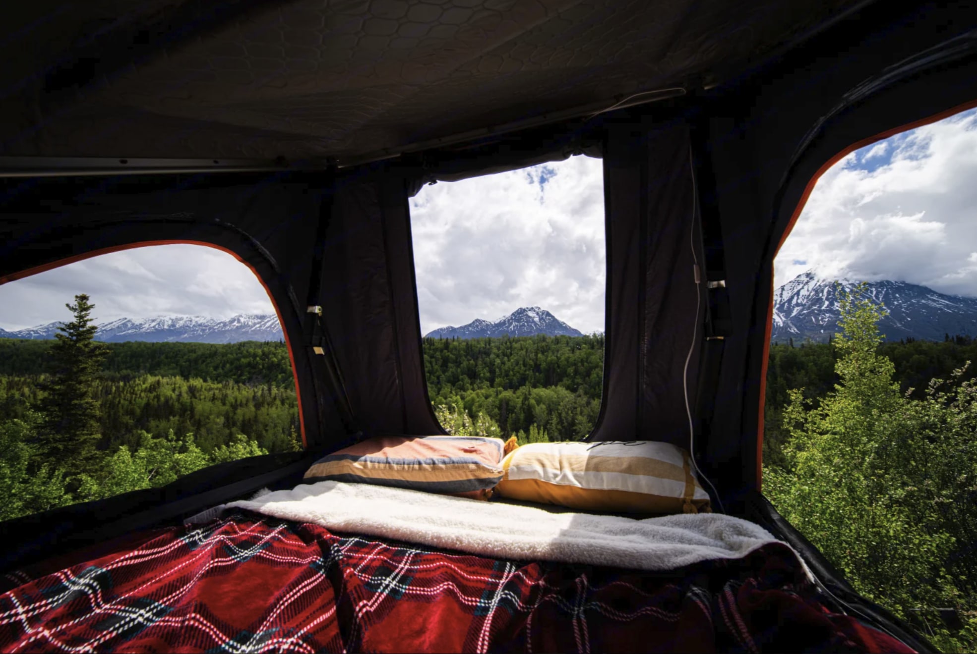 Cozy RV rental bed with pillows & plaid blanket, offering stunning views of green forests & snowy mountains through tent windows.. Toyota Highlander Roof Tent Camper 2015