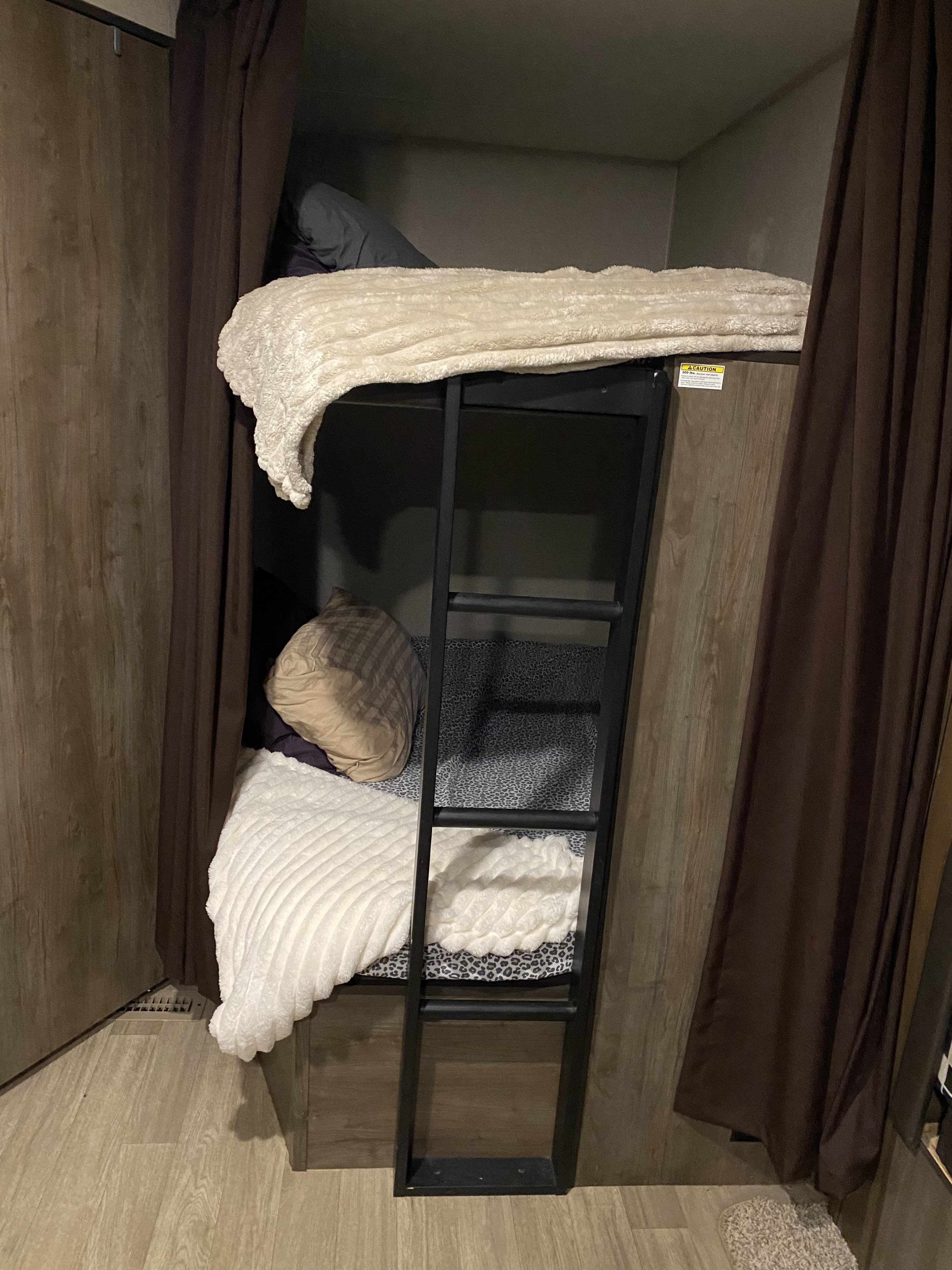 An RV rental's bunk beds, with a black ladder, soft white blankets, and brown privacy curtains.. Dutchmen Coleman Lantern 2018
