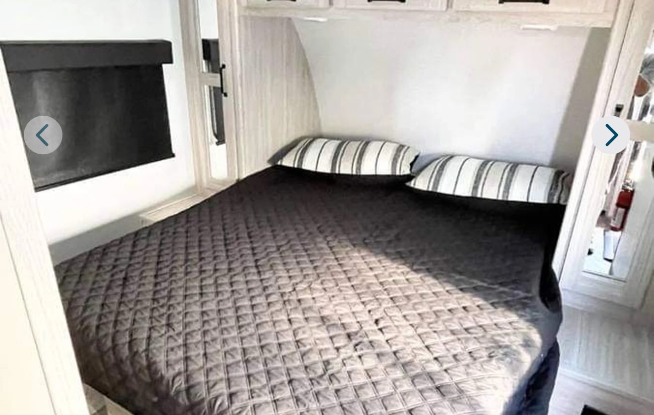 Inside a cozy RV rental bedroom: a quilted bed, striped pillows, overhead storage, and a window with a black shade.. Heartland RVs Sundance Ultra Lite 2021