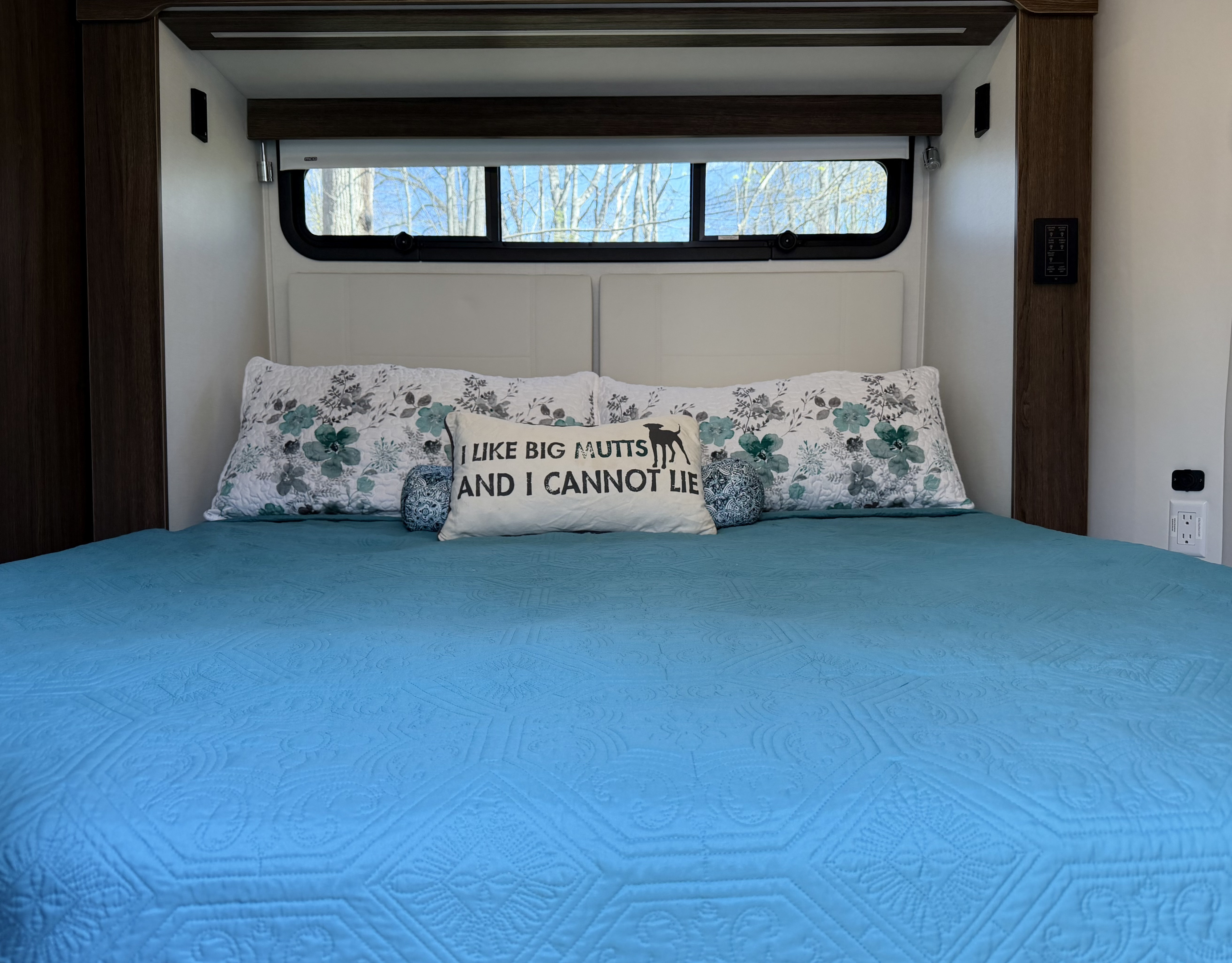 Cozy rv rental bedroom features a blue quilted bed, floral pillows, and a dog-themed pillow. Trees visible through the window.. Leisure Travel Vans Unity 2023