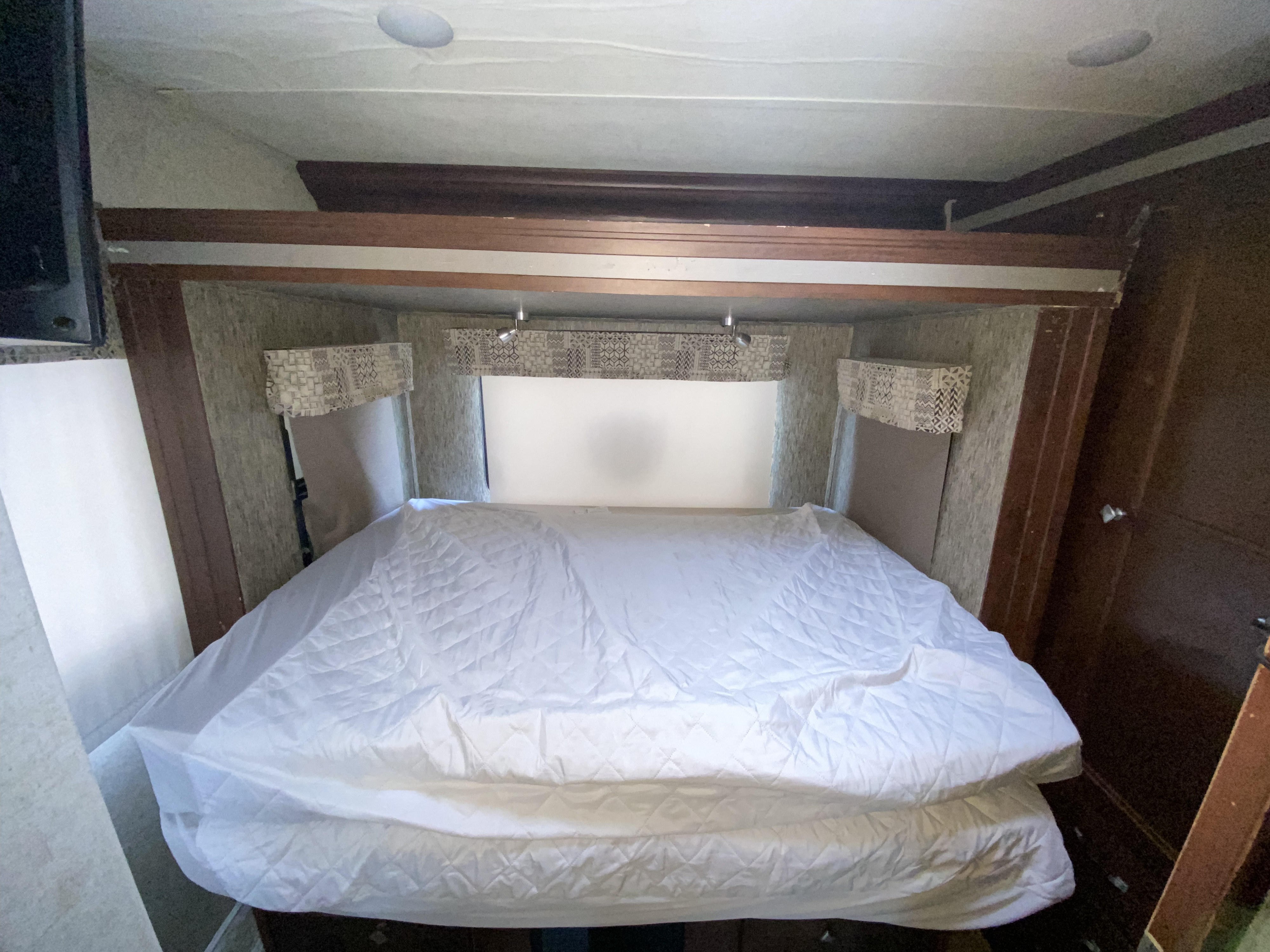 RV rental bedroom with a white-covered bed, framed by wood. A window with valances and lights is above the bed. TV on left, door on right.. Forest River Sunseeker 2017