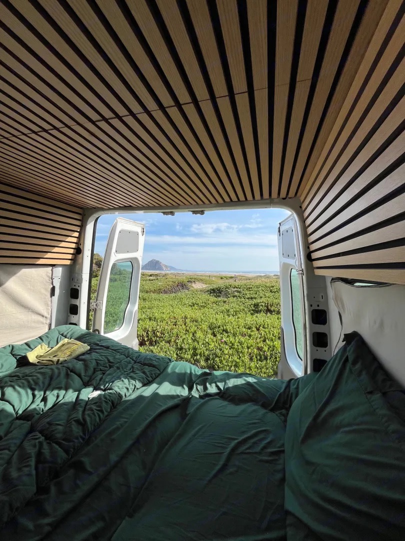 Comfortable RV rental campervan interior with green bed & wood slats, offering a scenic view of lush fields & ocean.. Mercedes-Benz Sprinter 2008