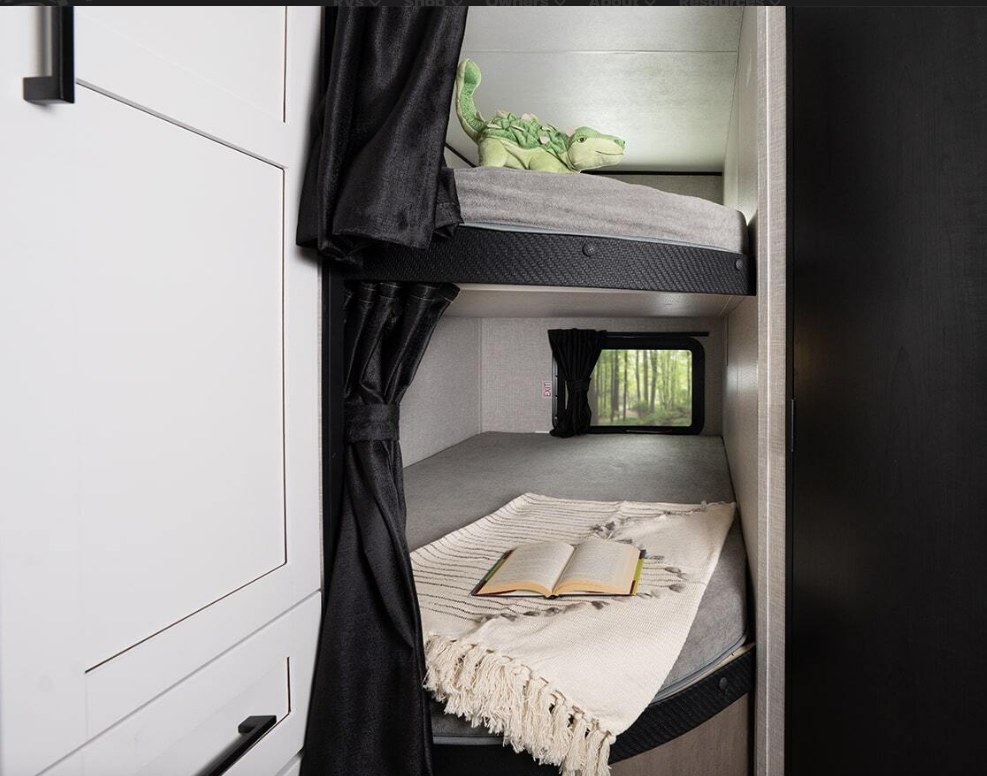 Cozy RV rental bunk beds with a window view of trees, a plush dinosaur, and a book on a blanket. Perfect for family travel.. Highland Ridge RV Open Range 2025