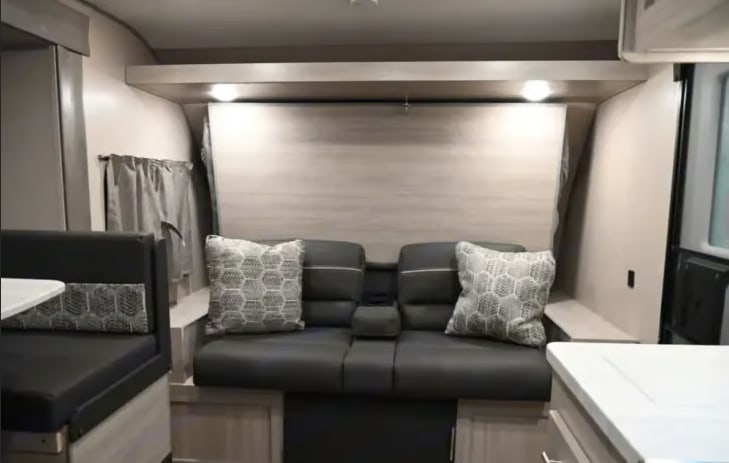 RV rental interior: Compact living space with a gray sofa, patterned pillows, a dinette on the left, and overhead lighting.. Coachmen Catalina Summit 2022