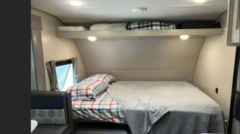 Inside an RV rental: a compact bedroom with a plaid bed, cozy grey blanket, overhead storage, and a window.. Coachmen Catalina Summit 2022